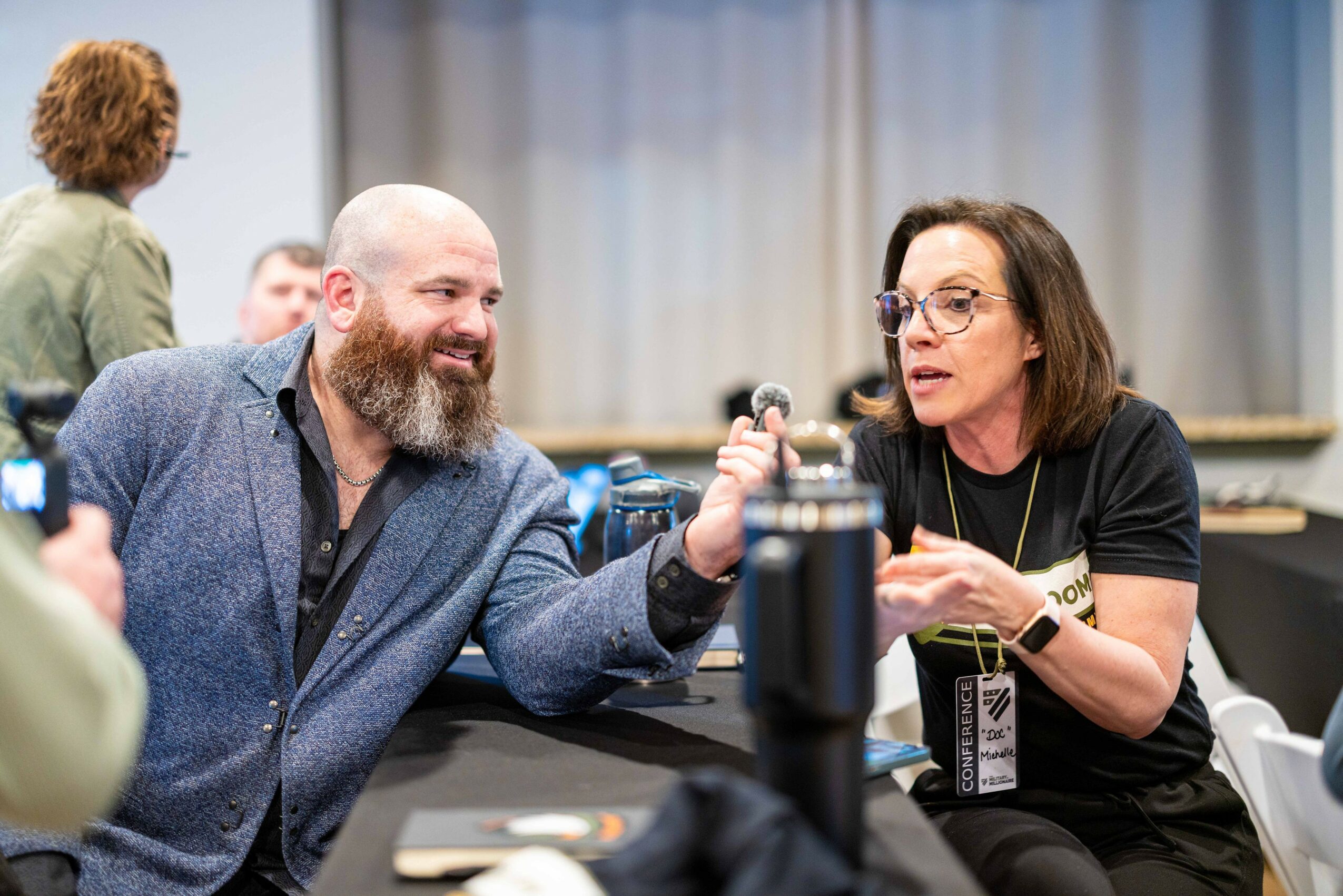 Two people engaged in conversation, one smiling and holding a microphone, in a room with others in the background.