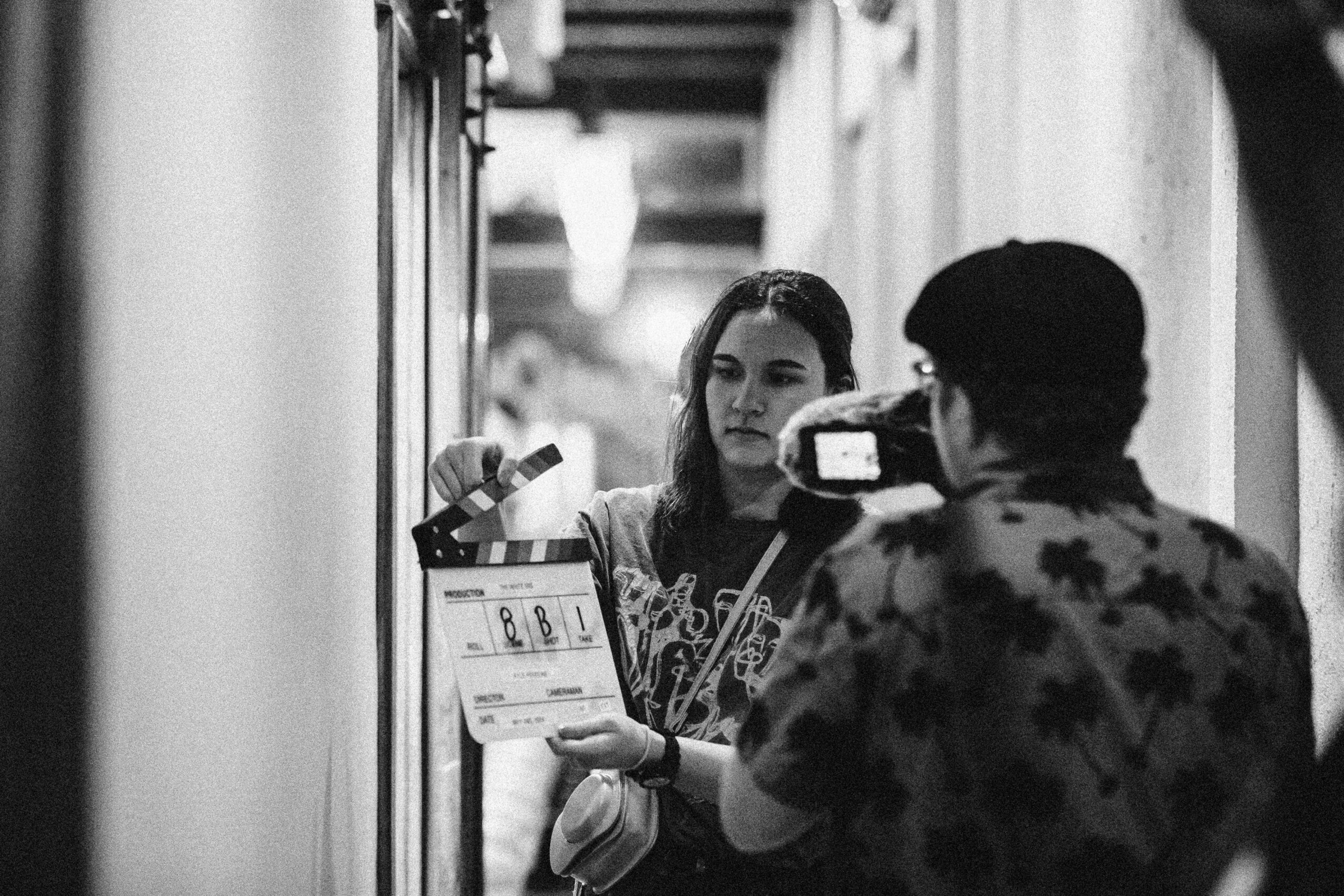 Person holding a clapperboard filming a woman in a narrow corridor, black and white image.