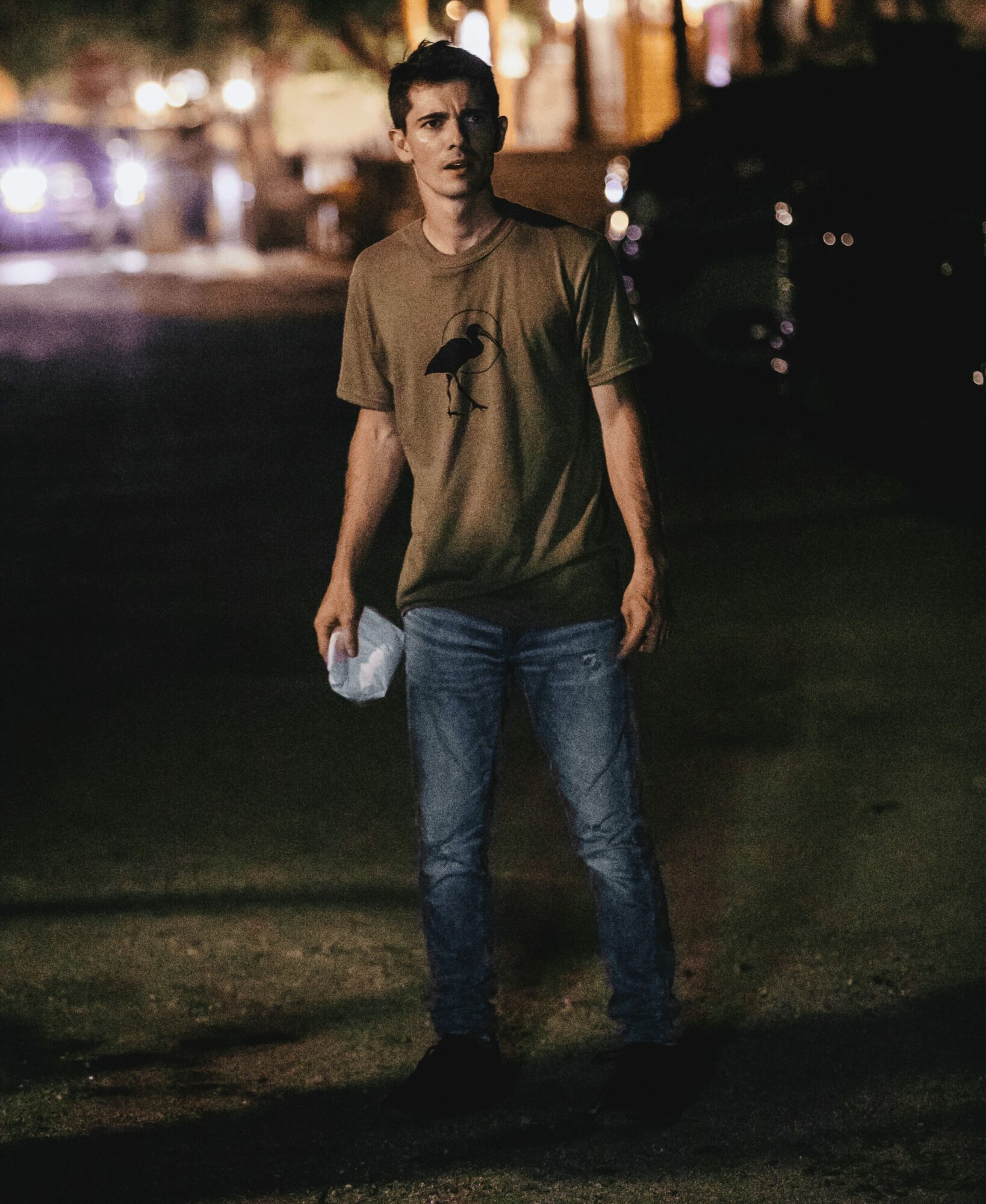 Young man standing outdoors at night, holding a plastic bag, with blurred city lights in background.