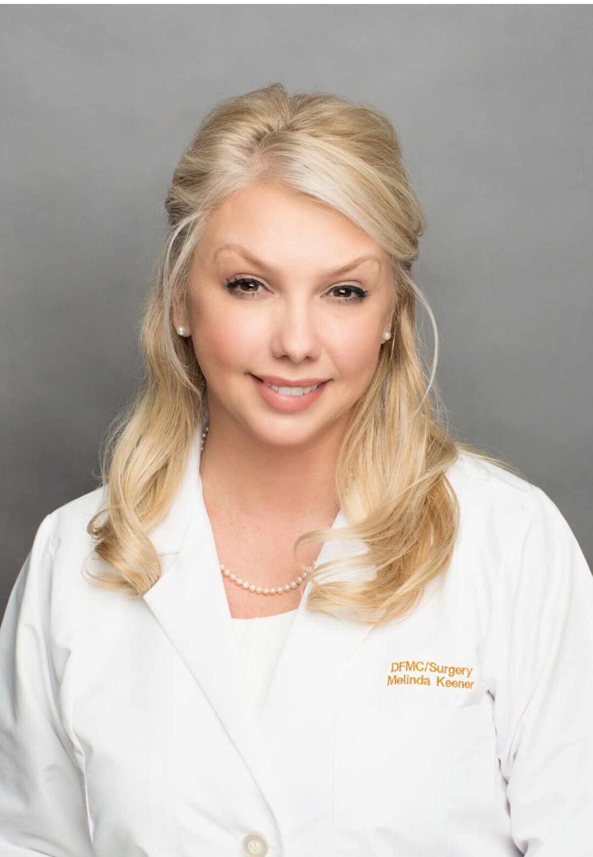 Portrait of a woman with blonde hair, smiling, wearing a white coat with a name tag, against a gray background.