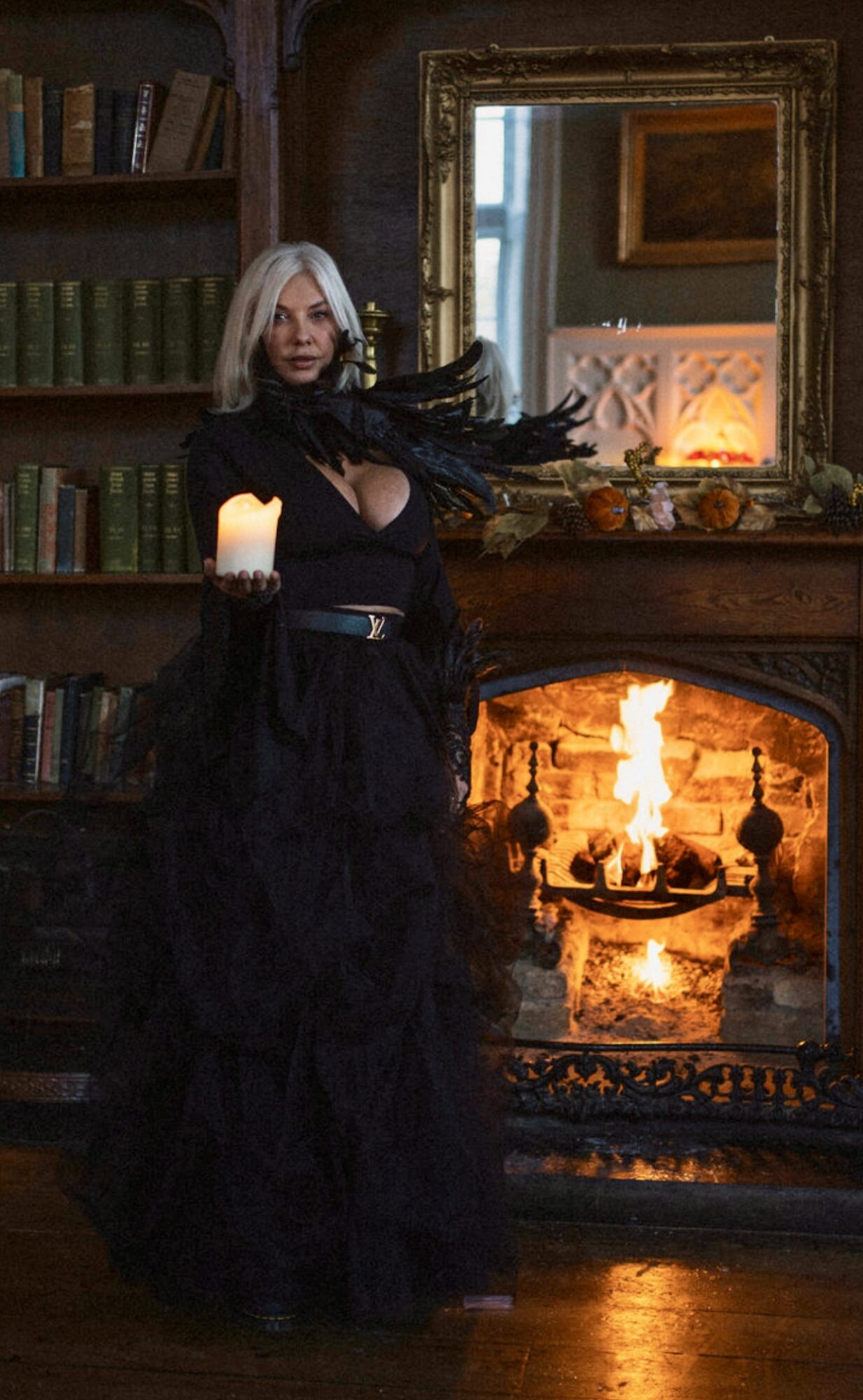 Woman with long white hair standing in front of a fireplace with a mirror above, holding a candle, in a dimly lit room.