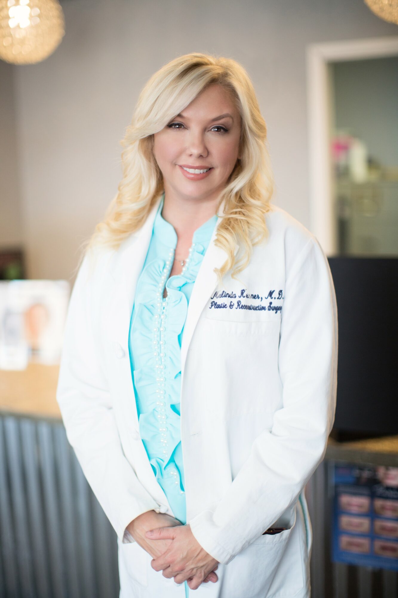 Woman with blonde hair in a white coat and light blue blouse, smiling in a professional setting.