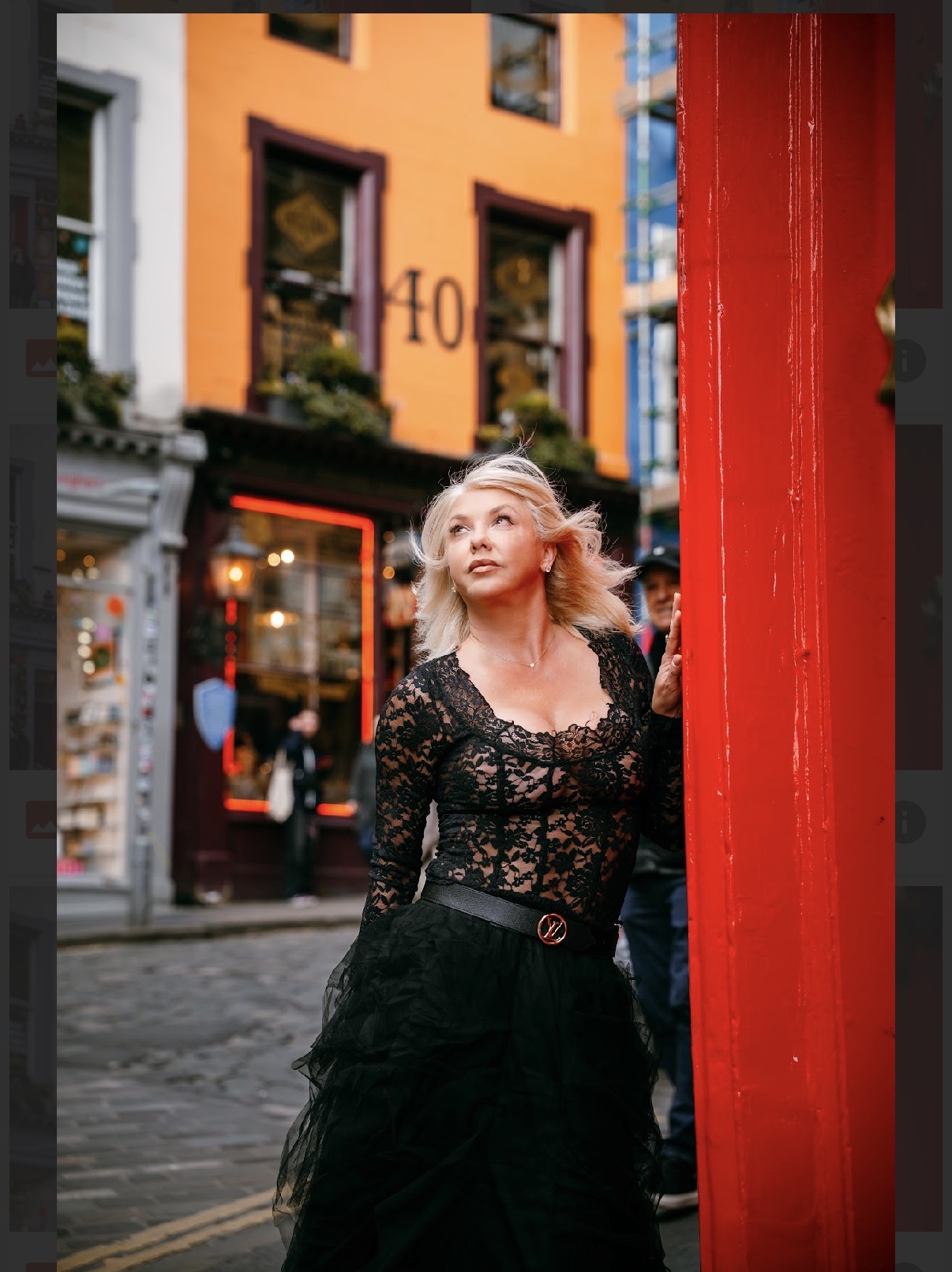 Woman with blonde hair in black lace dress standing near a red pole on a city street, colorful buildings in background.