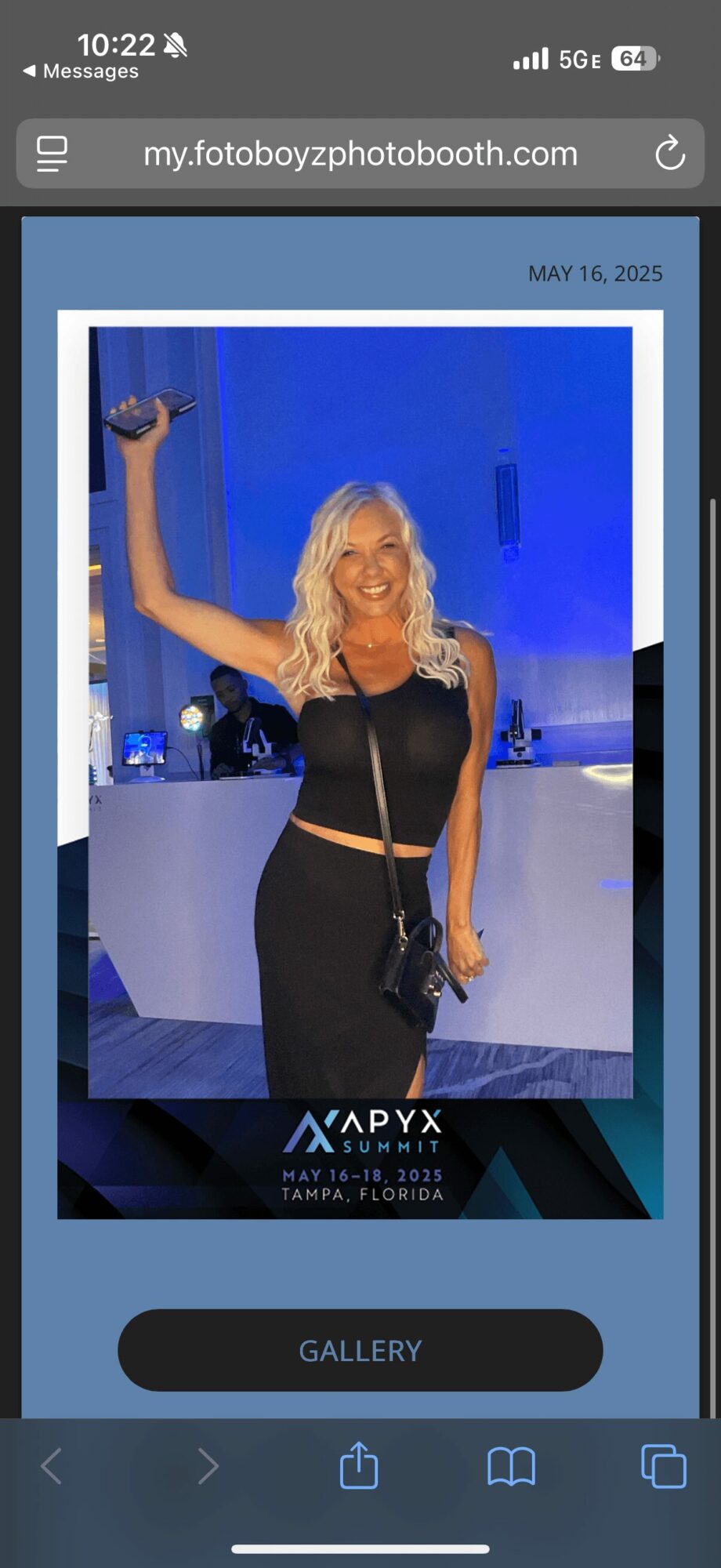 Woman with blonde hair in black dress smiling and waving at event, blue background, holding a small bag.