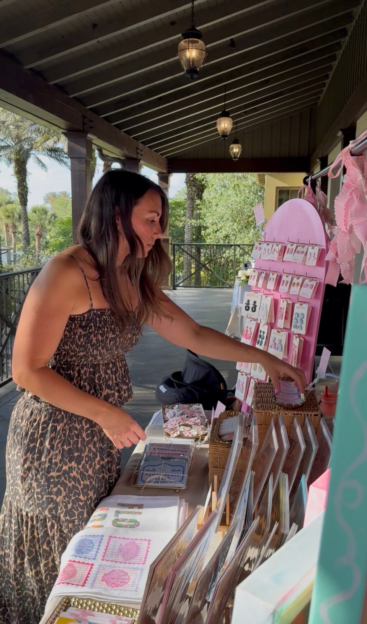 Woman in leopard print dress shopping at an outdoor kiosk with pink accessories, under a covered area with hanging lights.