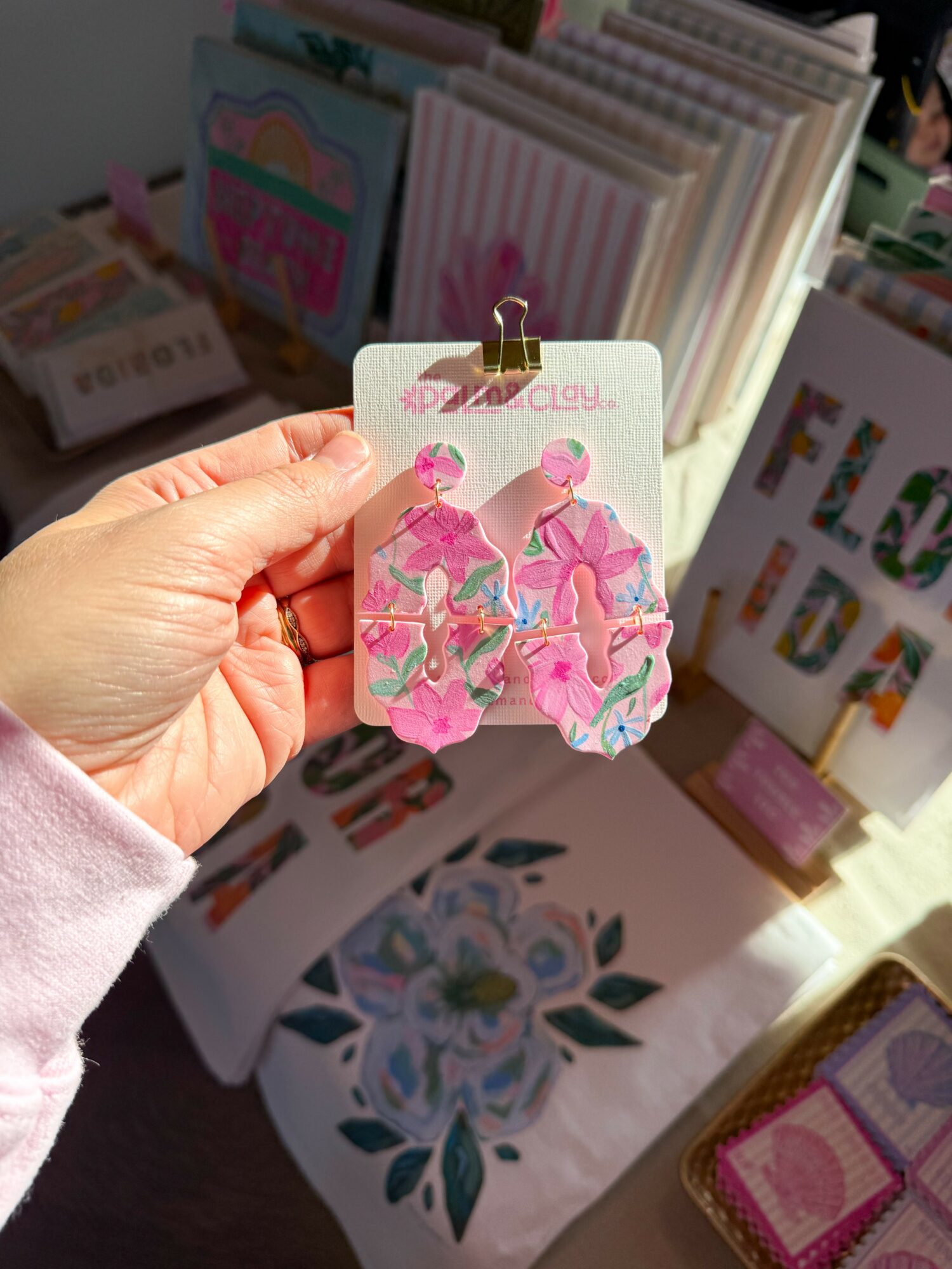 Hand holding pink flamingo-shaped earrings with a rainbow background, surrounded by colorful cards and decorations.