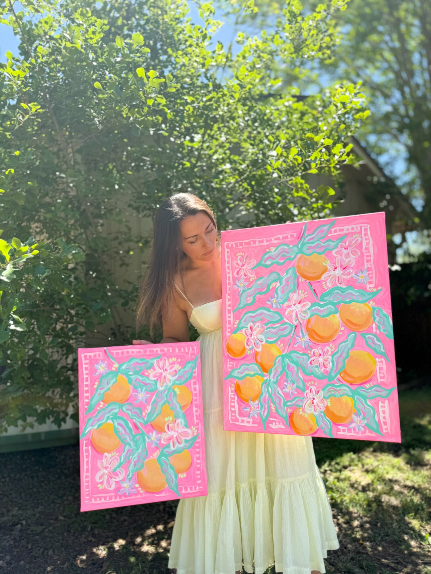Young woman in a light-colored dress holding two colorful paintings outdoors with green trees in background.
