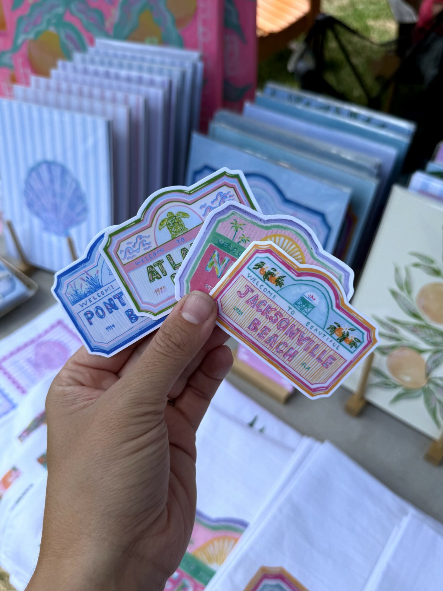 Hand holding colorful paper tickets with beach and rainbow designs, displayed at an outdoor market stall.