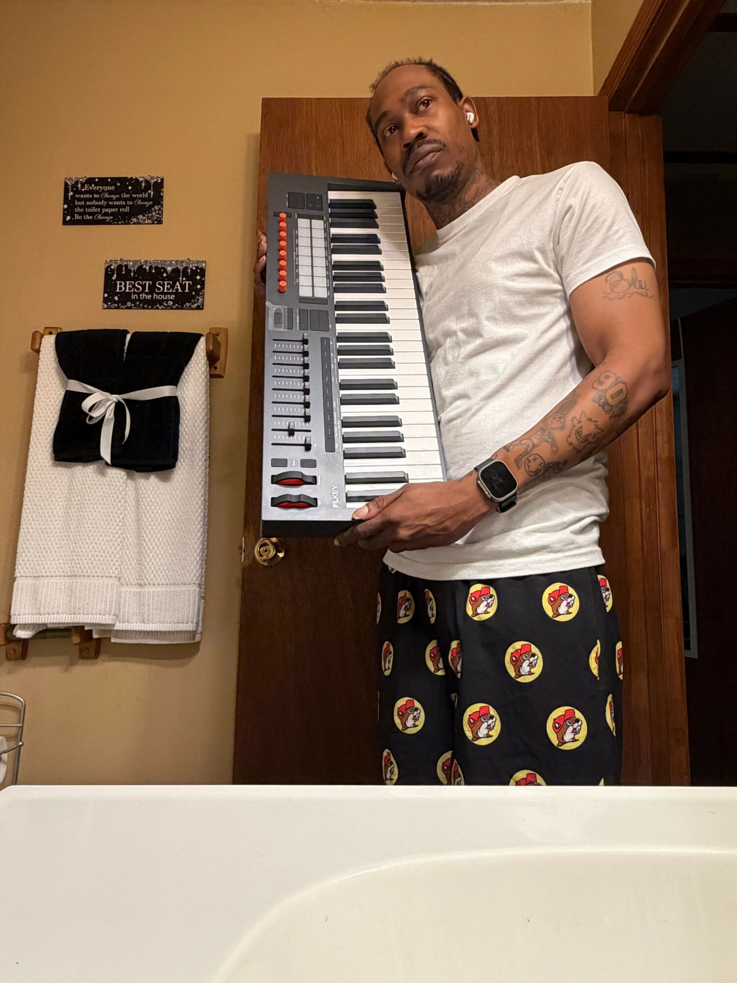 Man standing in a room holding a keyboard, wearing a white t-shirt and shorts with smiley face patterns, with a towel on a rack behind him.