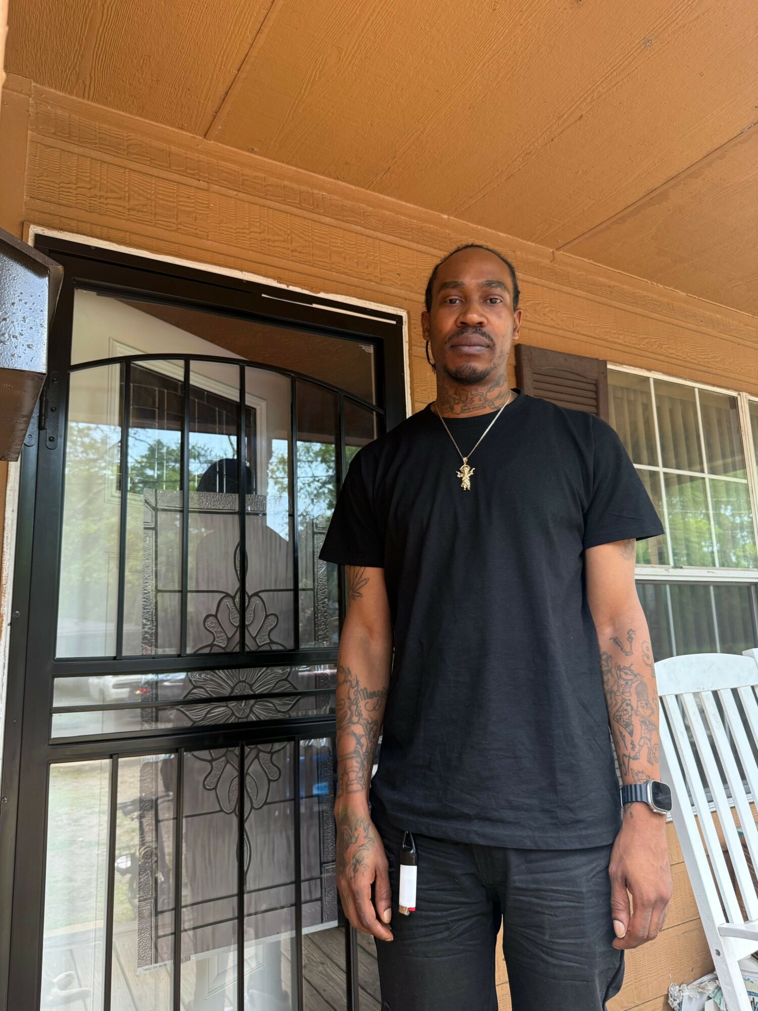 Man standing indoors near a window with a wooden frame, wearing a black t-shirt and watch, holding a phone.