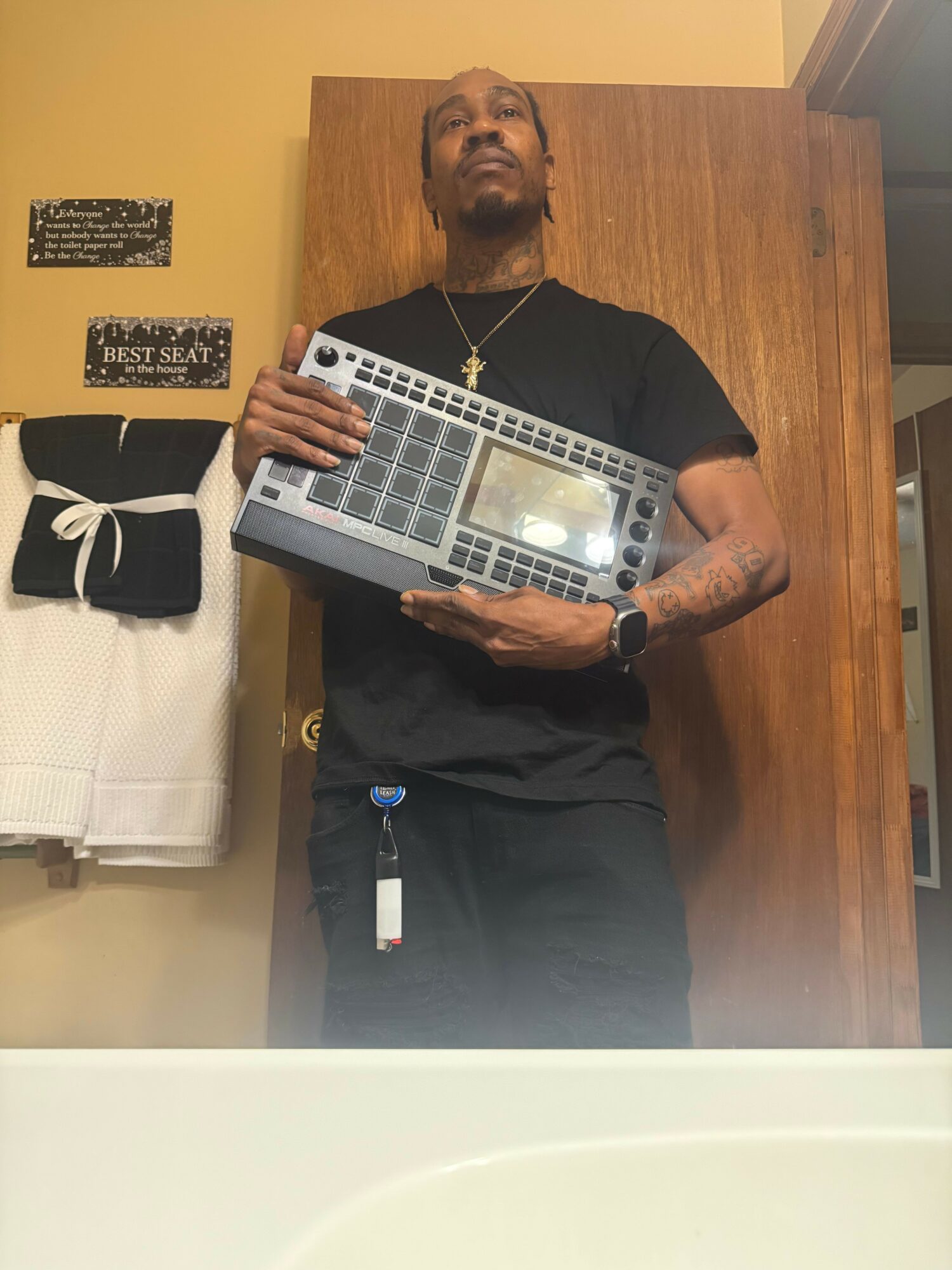Man with tattoos holding a laptop, standing in front of a wooden wall and a white towel, wearing a black shirt and pants.