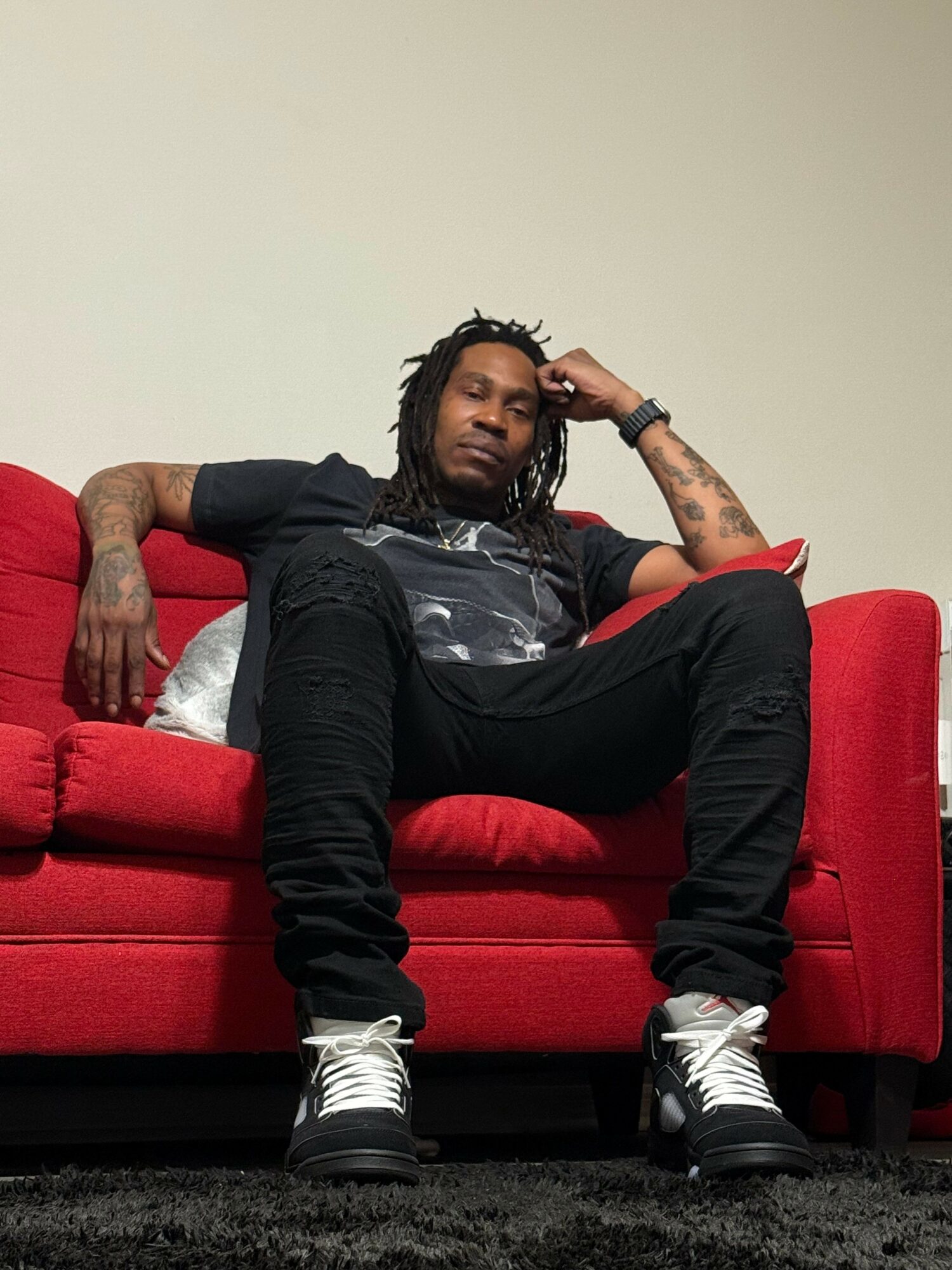 Person with tattoos sitting on a red couch, wearing black pants, a black shirt, and white sneakers, with a neutral background.