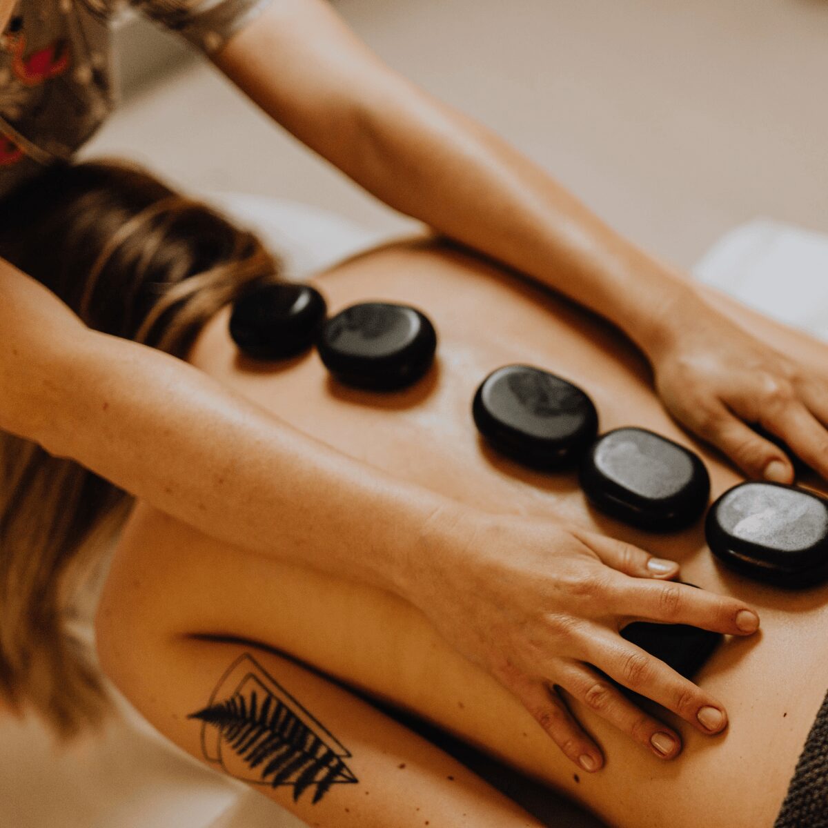 Person receiving a massage with hot stones on back, tattoo visible on shoulder, massage therapist's hands applying pressure.