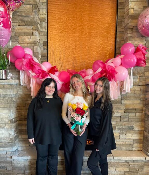 Three women standing in front of a decorated fireplace with pink balloons and a wooden panel, one holding a bouquet of flowers.