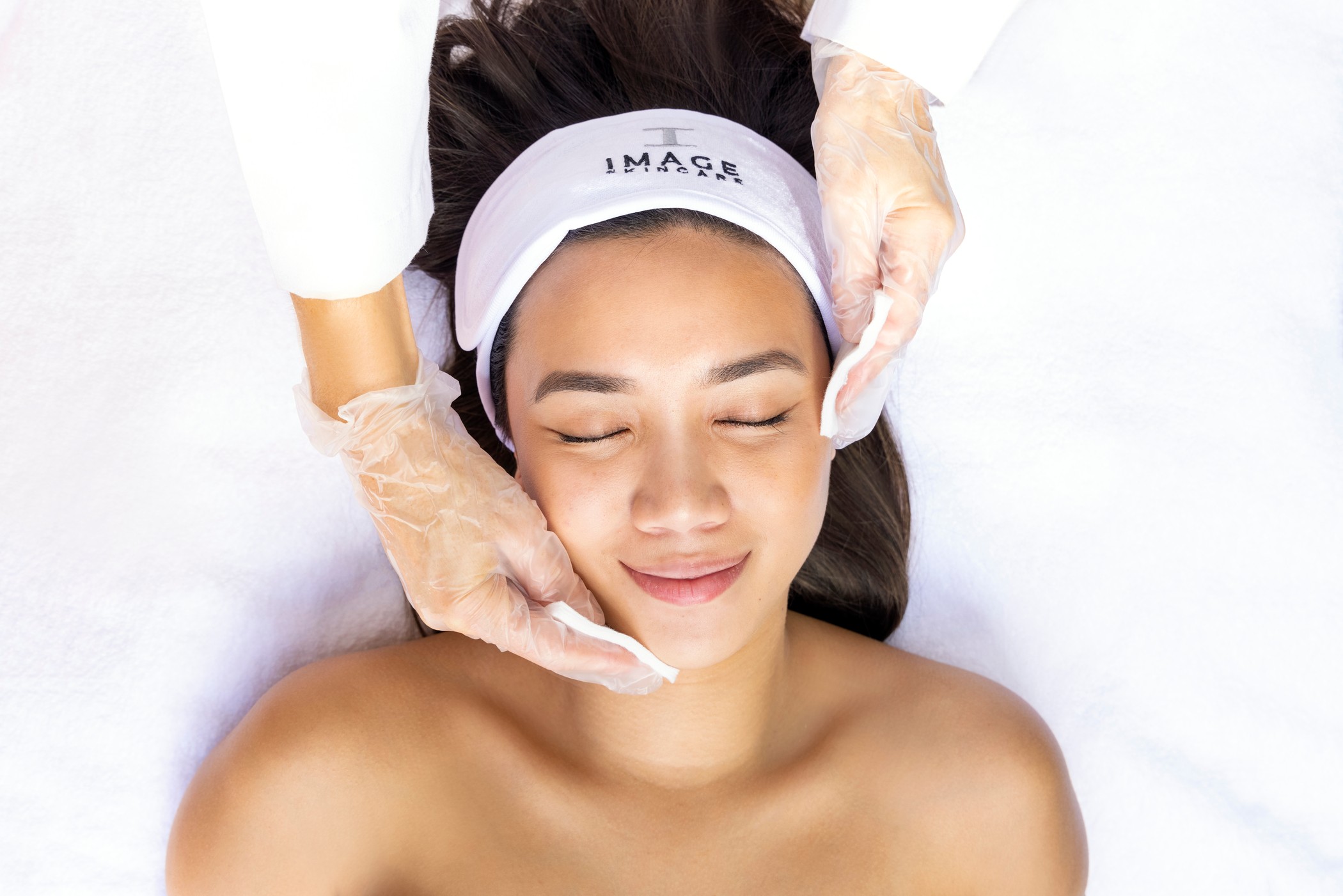 Woman receiving facial treatment with eyes closed, lying on a white surface, wearing a headband and gloves.