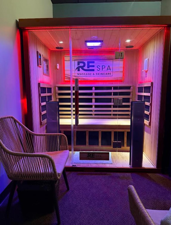 Small enclosed spa room with glass door, illuminated by red and blue lights, sign reads 'RE SPA MASSAGE & SKINCARE', chair outside