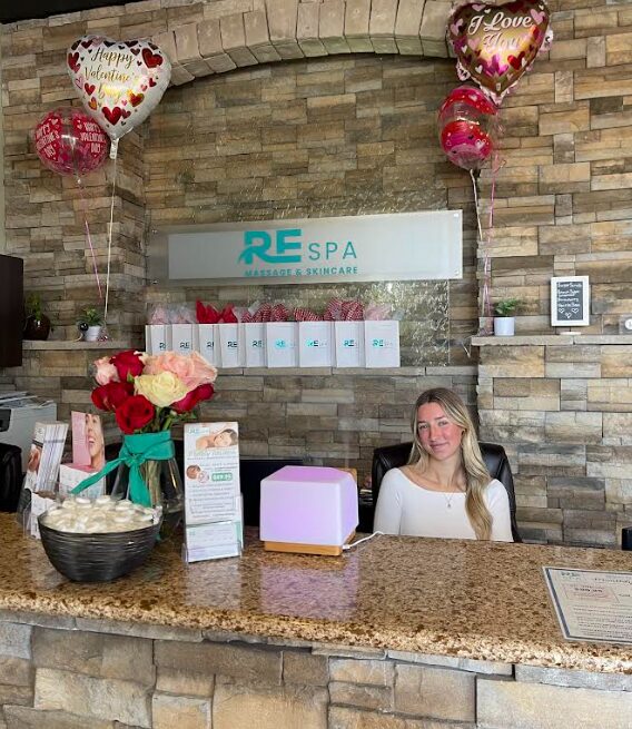Reception desk with a woman smiling, decorated with balloons and flowers, in a spa setting with a stone wall background.