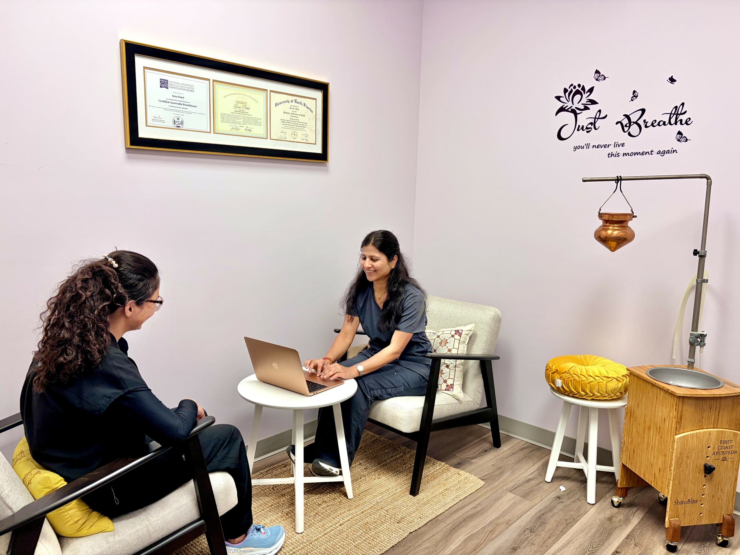 Two women sit in a room with certificates on the wall, one using a laptop, the other listening, with a small table between them.