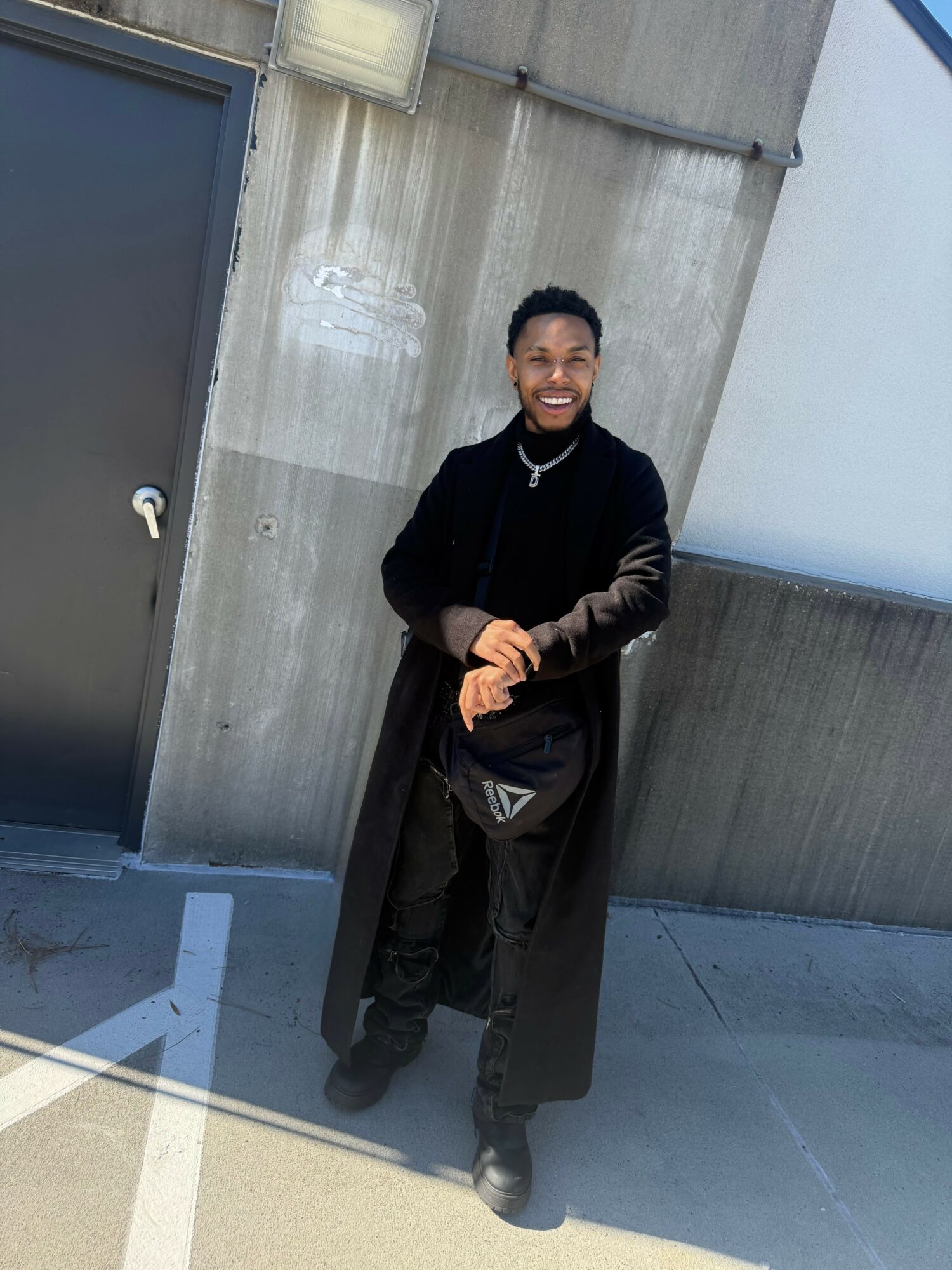Person standing outdoors against concrete wall, smiling, wearing black coat and pants, holding gloves, with sunlight on ground.