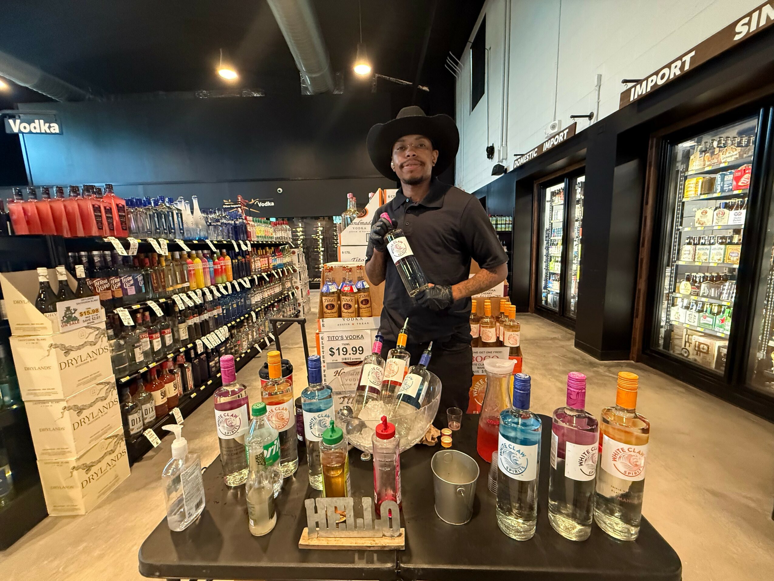 Person wearing a hat and black shirt stands behind a table with various bottles and cups in a store aisle.