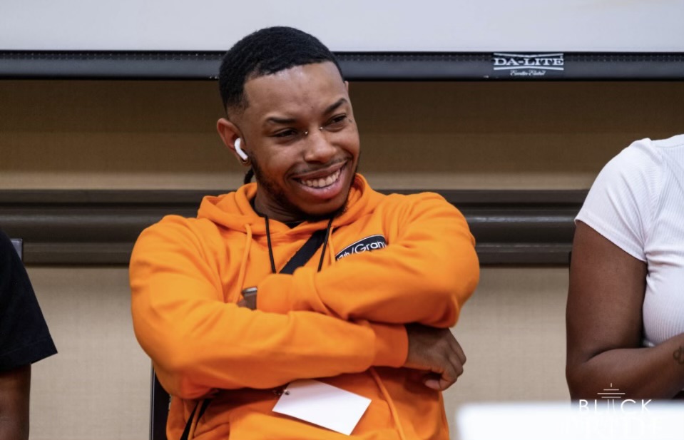 Smiling man with crossed arms wearing an orange hoodie, sitting in a classroom, with a blackboard behind him.