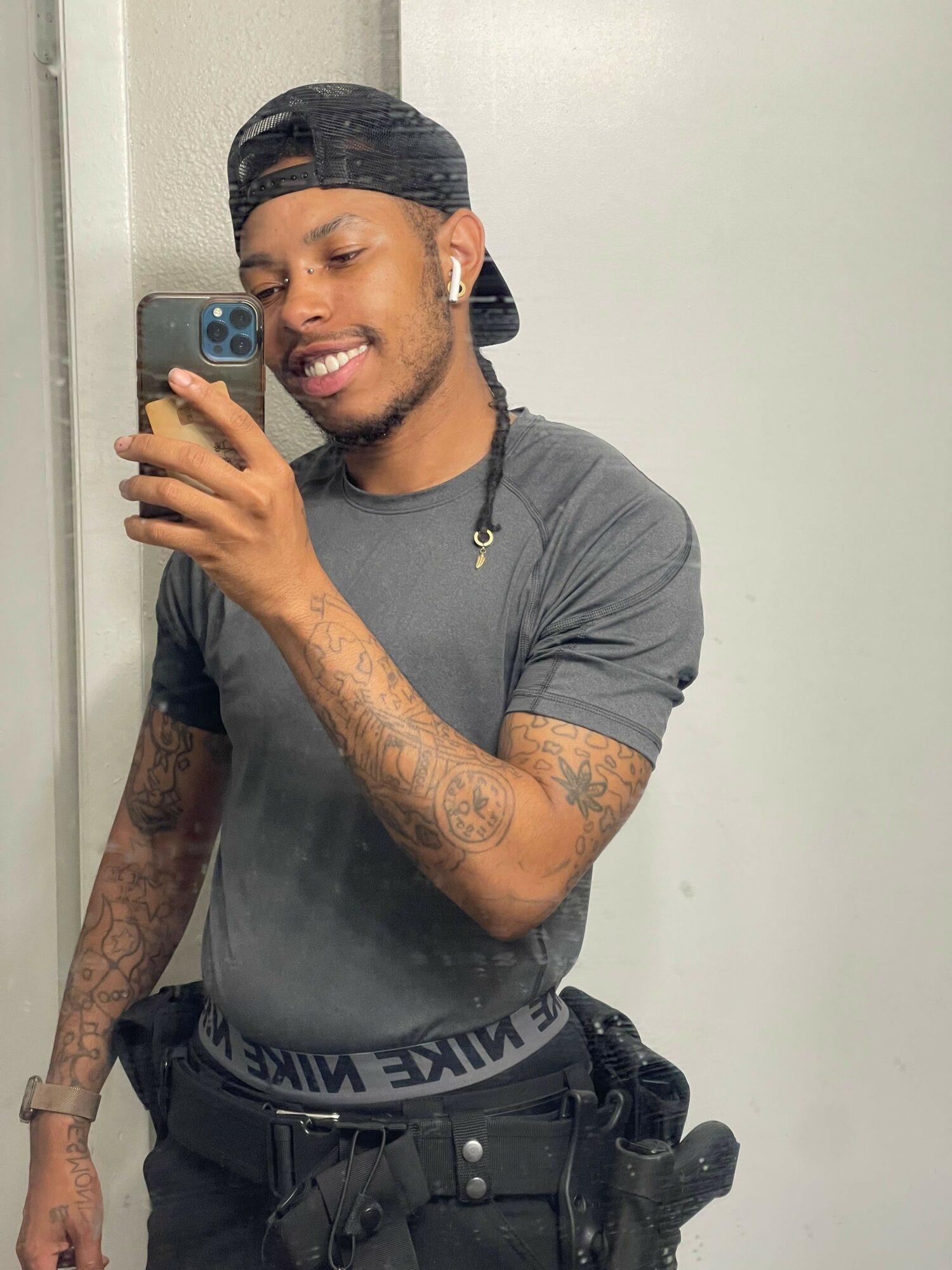 Young man taking a mirror selfie, smiling, wearing a gray t-shirt, black cap, and a black waist bag, with tattoos on arms.