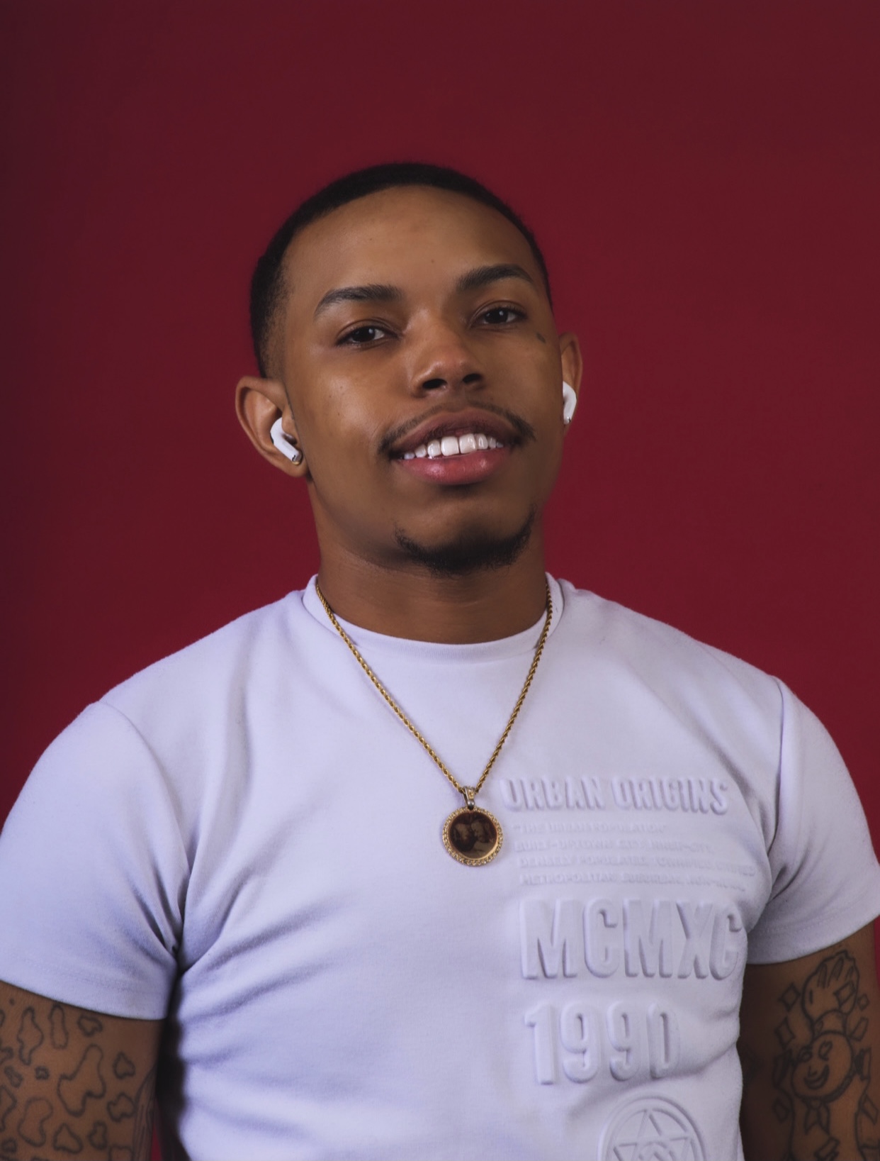 Young man with short hair, wearing a white T-shirt, gold necklace, and earrings, smiling against a red background.