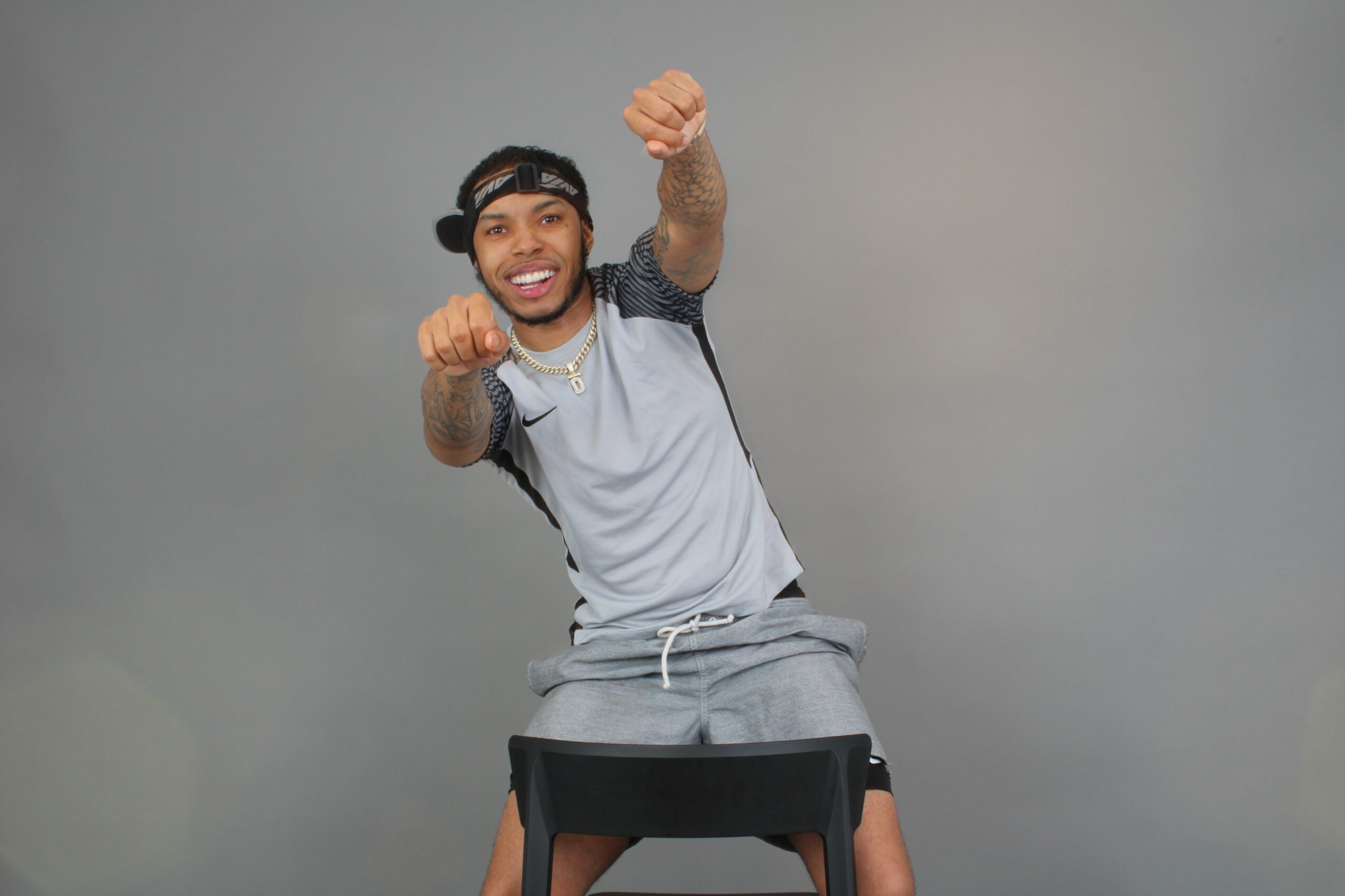 Young man smiling and making a fist gesture, sitting on a black chair against a plain gray background.