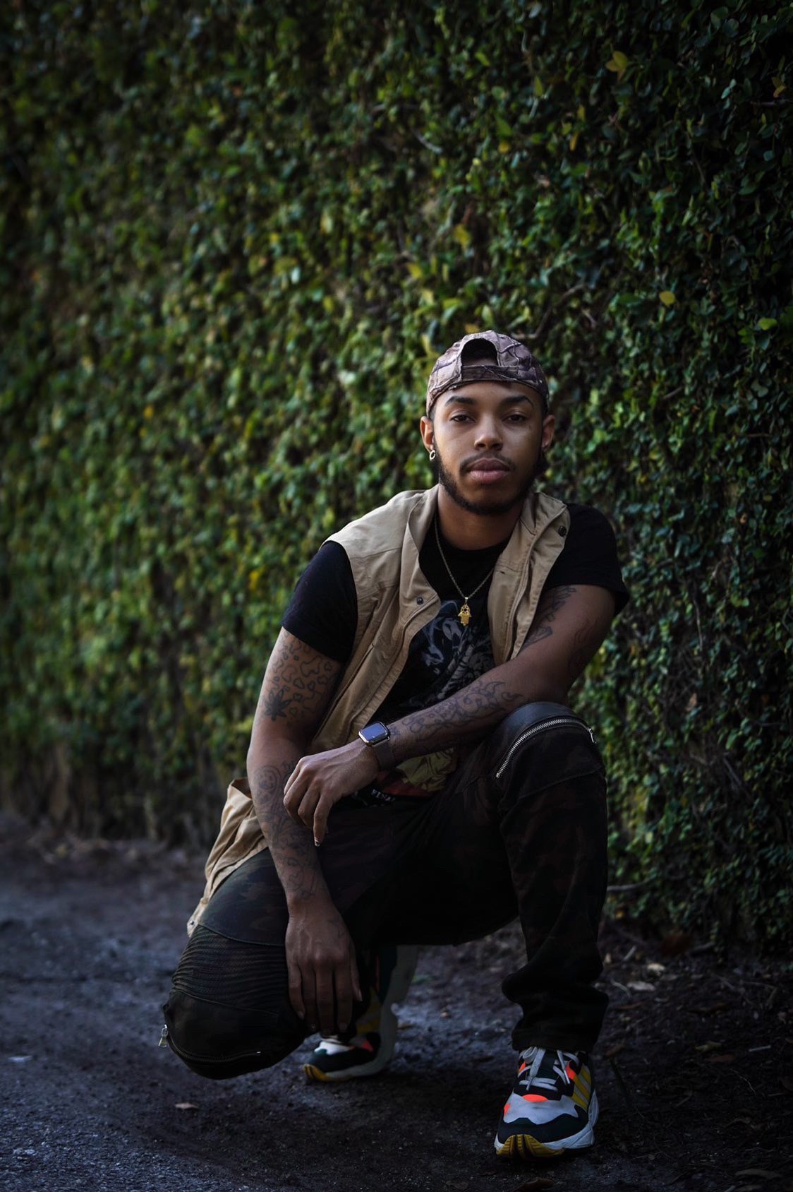Young man crouching outdoors in front of a green hedge, wearing a cap, black shirt, and sneakers.