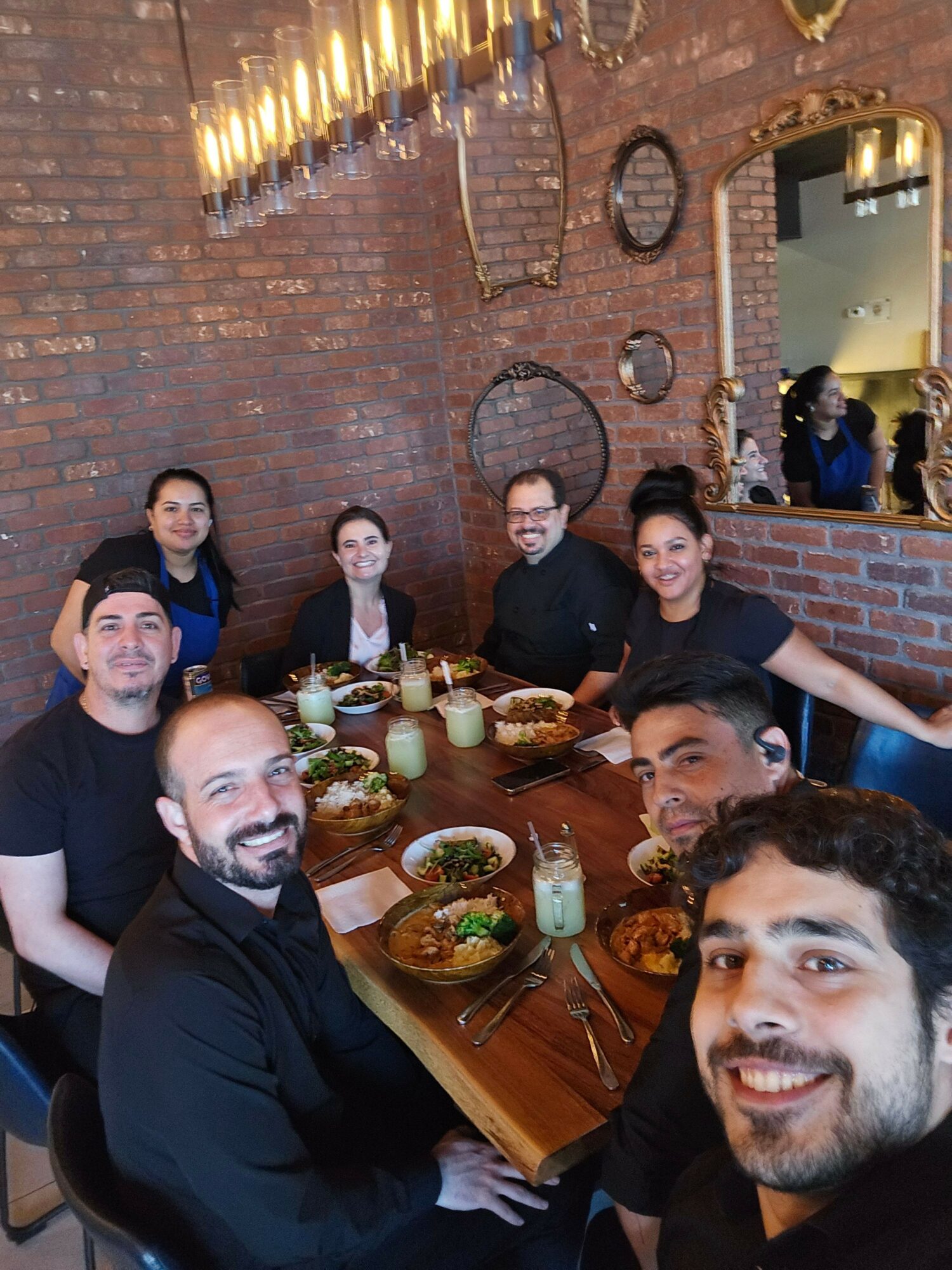 Group of people smiling and enjoying a meal at a table in a brick-walled restaurant.