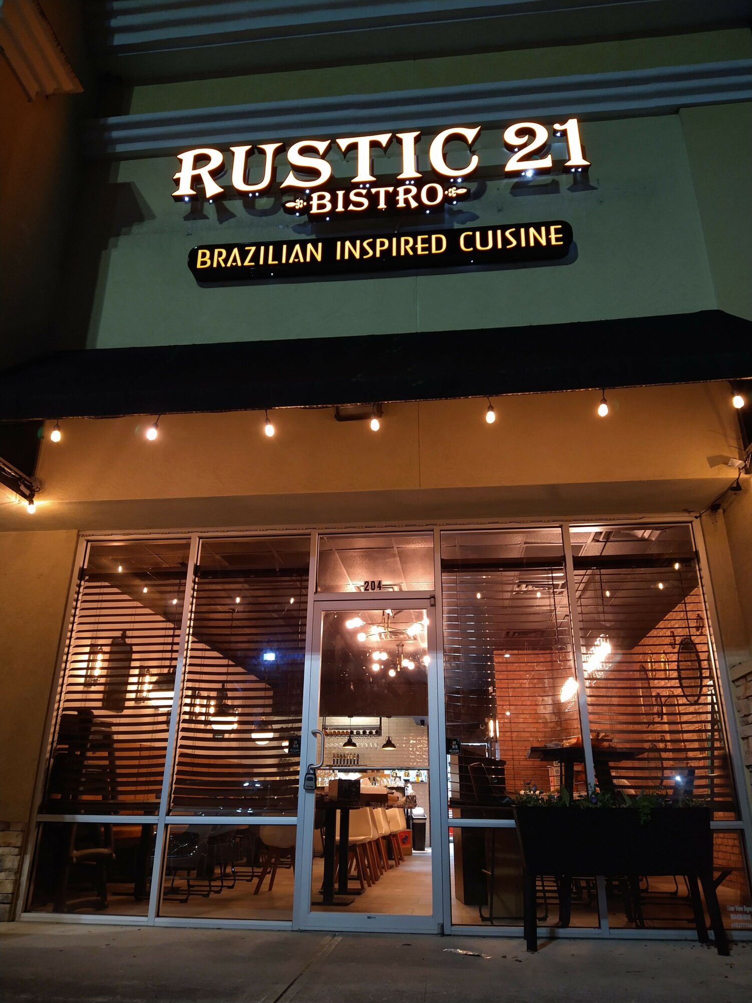 Restaurant interior with warm lighting, tables, and chairs visible through large glass windows, viewed from outside at night.