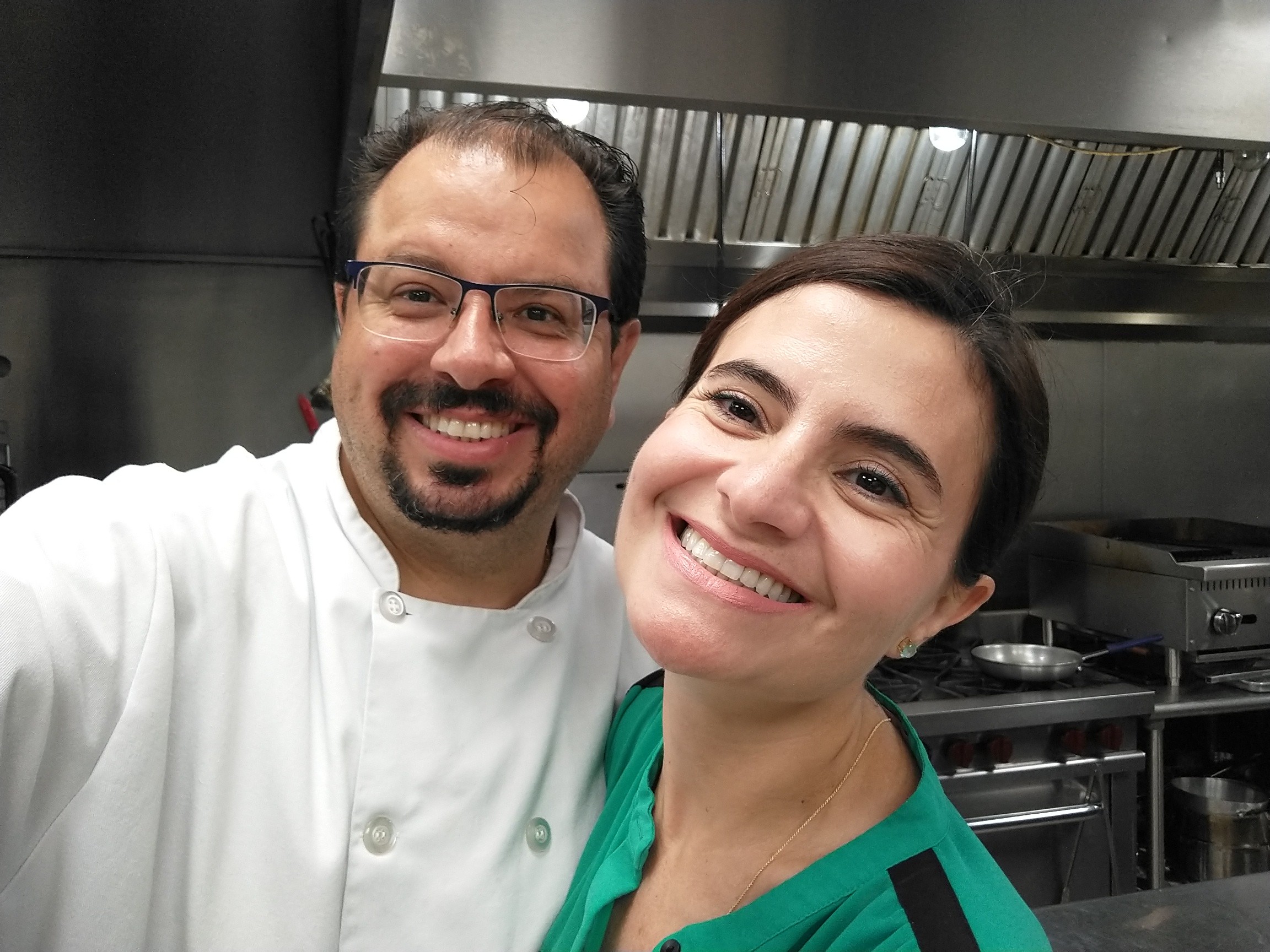 Two people smiling in a kitchen, one wearing a white chef's coat, the other in a green top.