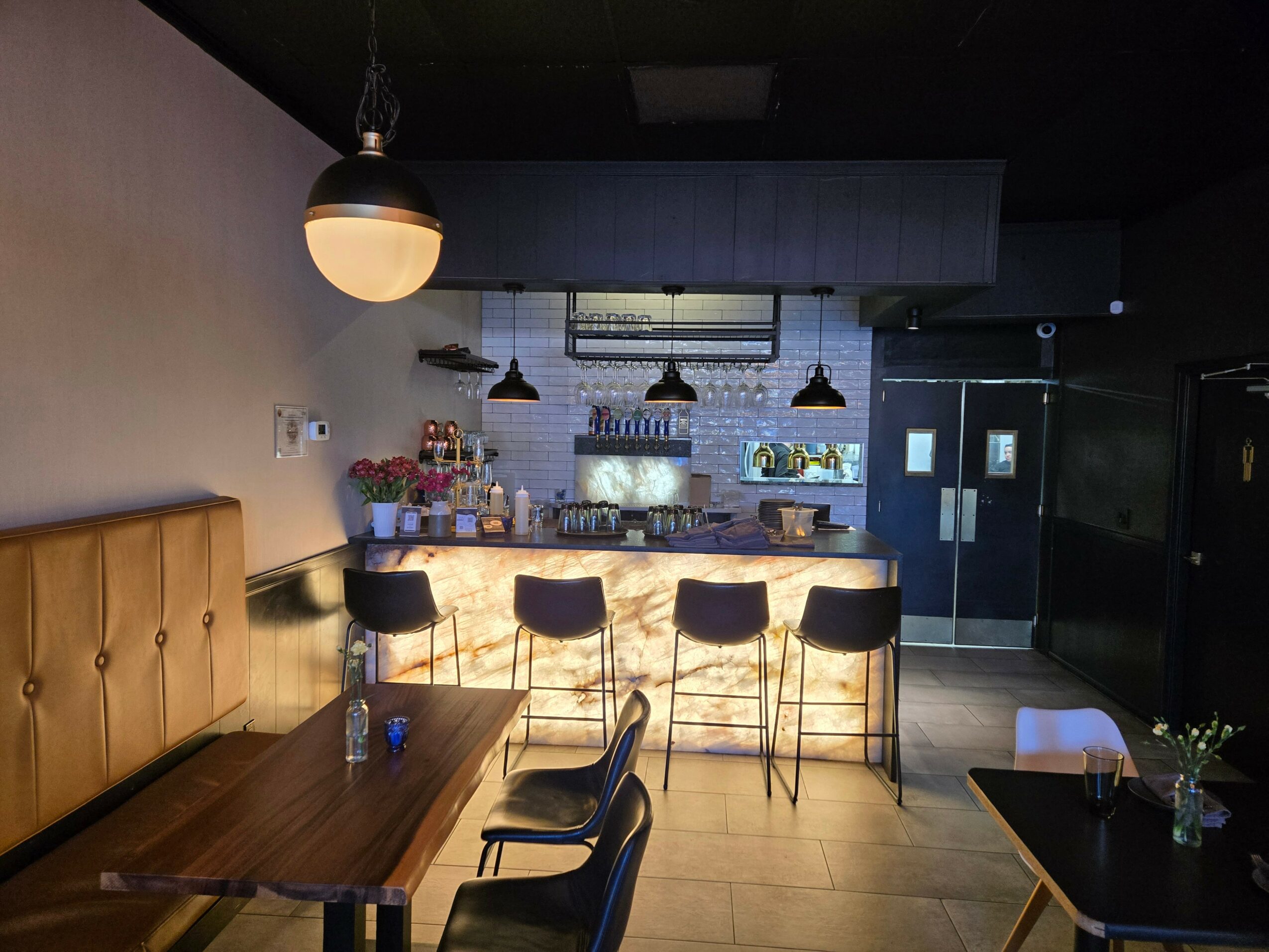 Bar area with four black stools, a wooden table with chairs, and a counter with bottles and glasses, dim lighting.
