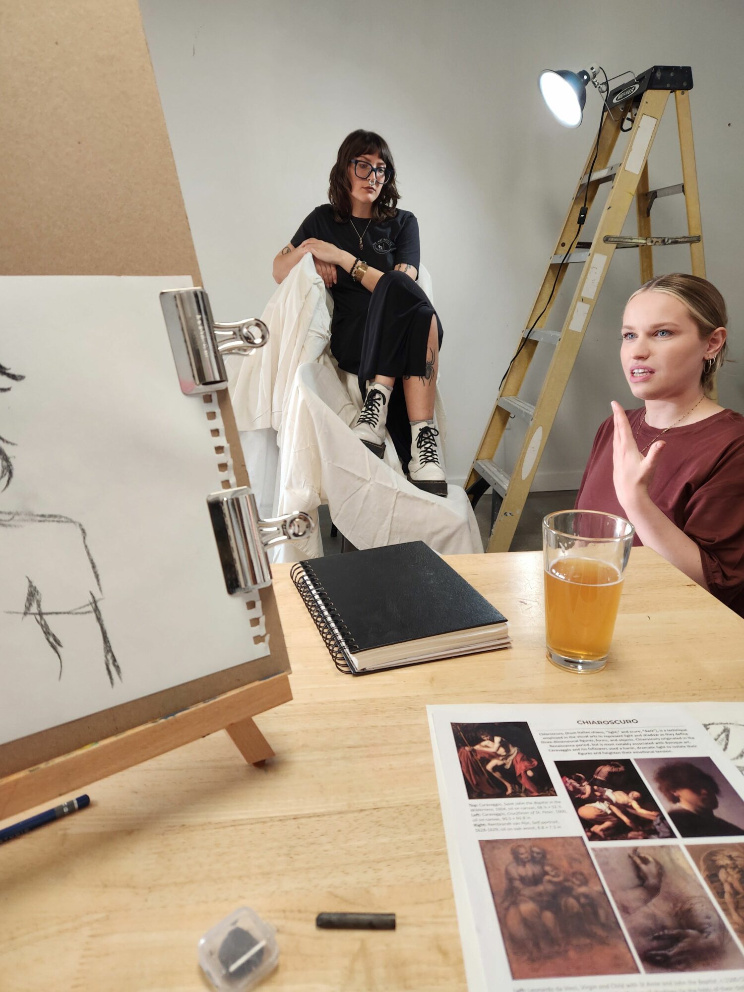 Two women sit at a table with a notebook, a glass of beer, and printed images, in a room with a ladder and lighting equipment.