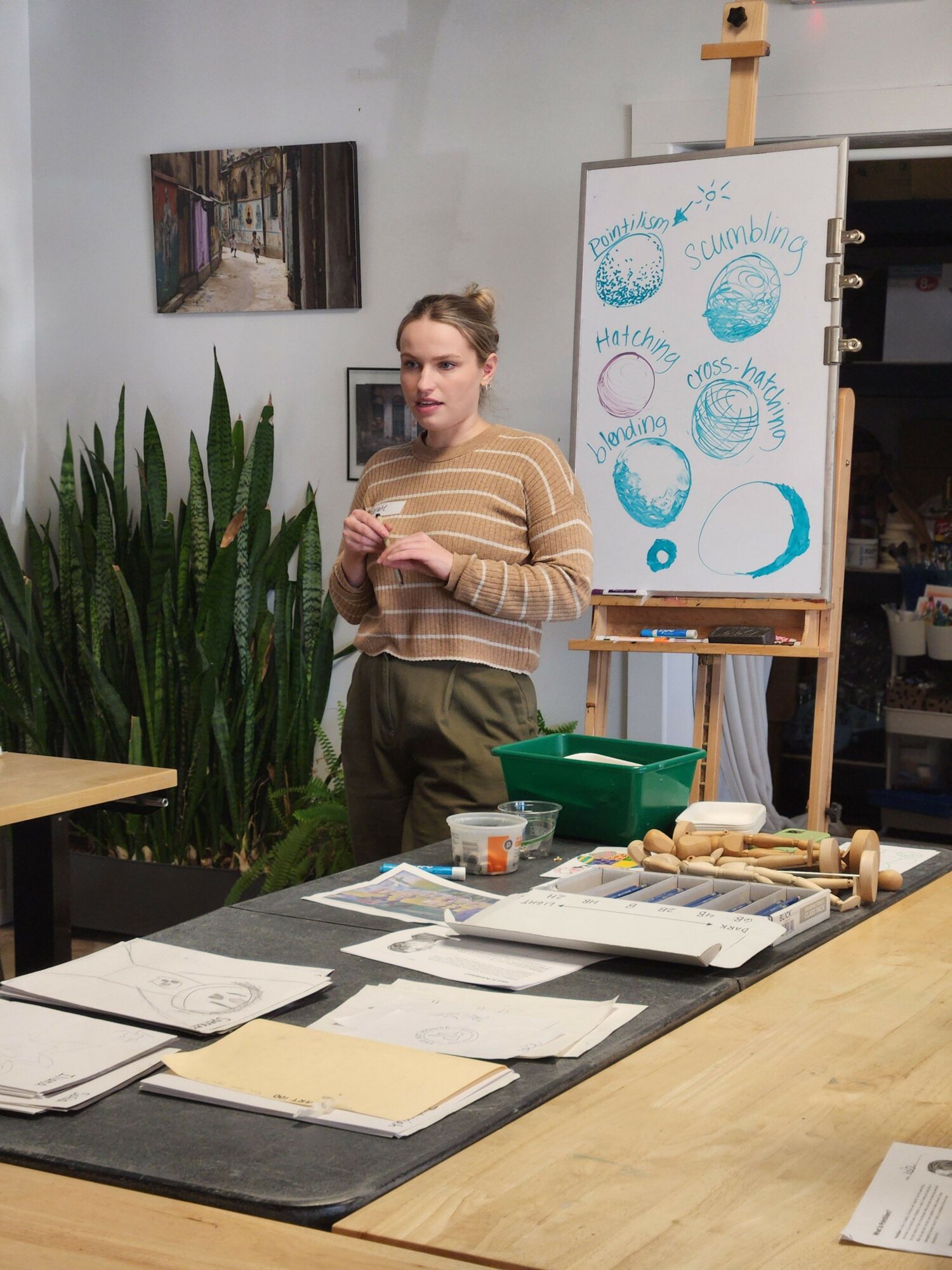 Woman standing near a table with papers and a whiteboard with drawings, in a room with plants and artwork.