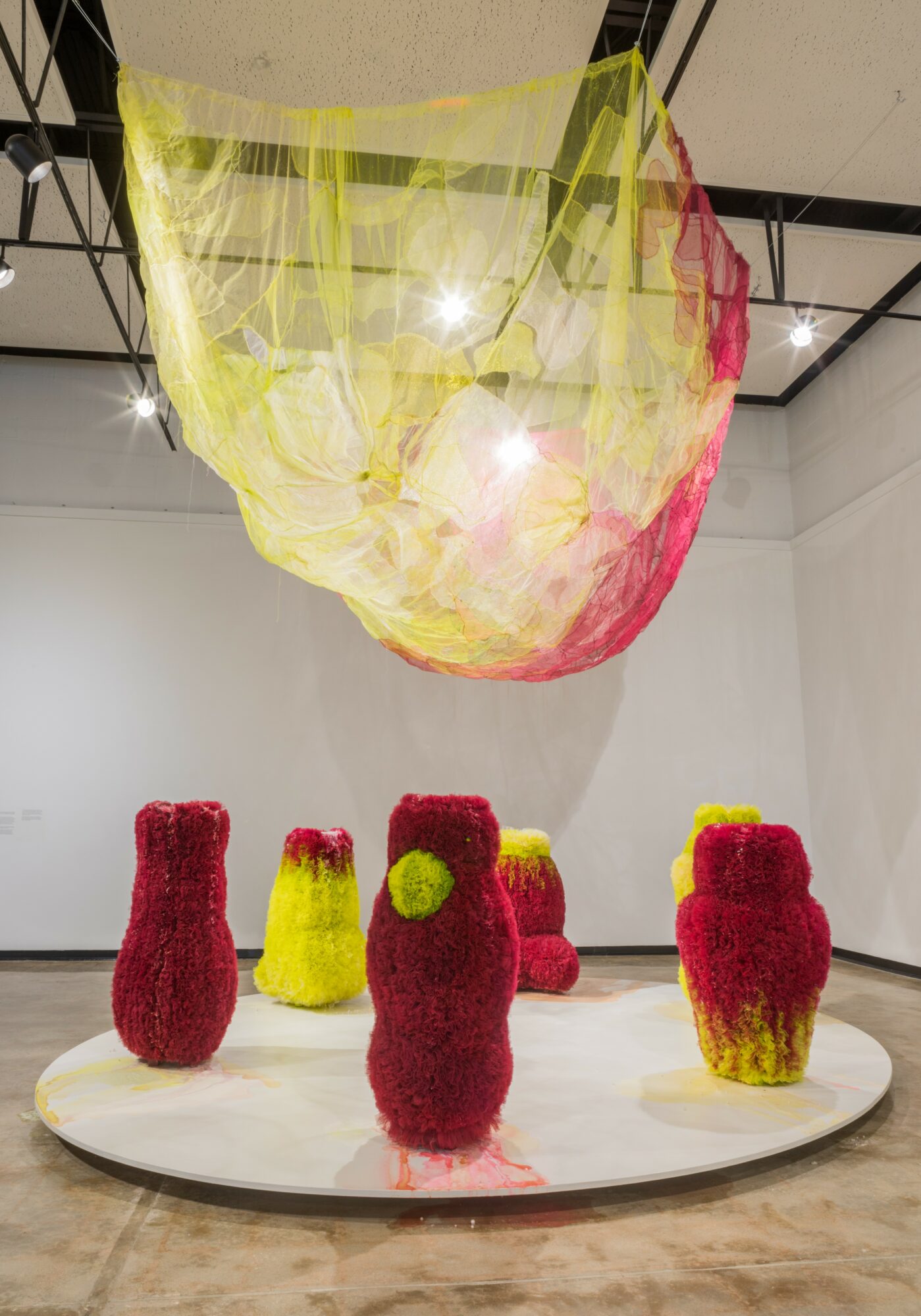 Colorful fabric sculptures resembling abstract figures on a white circular platform in a gallery space.