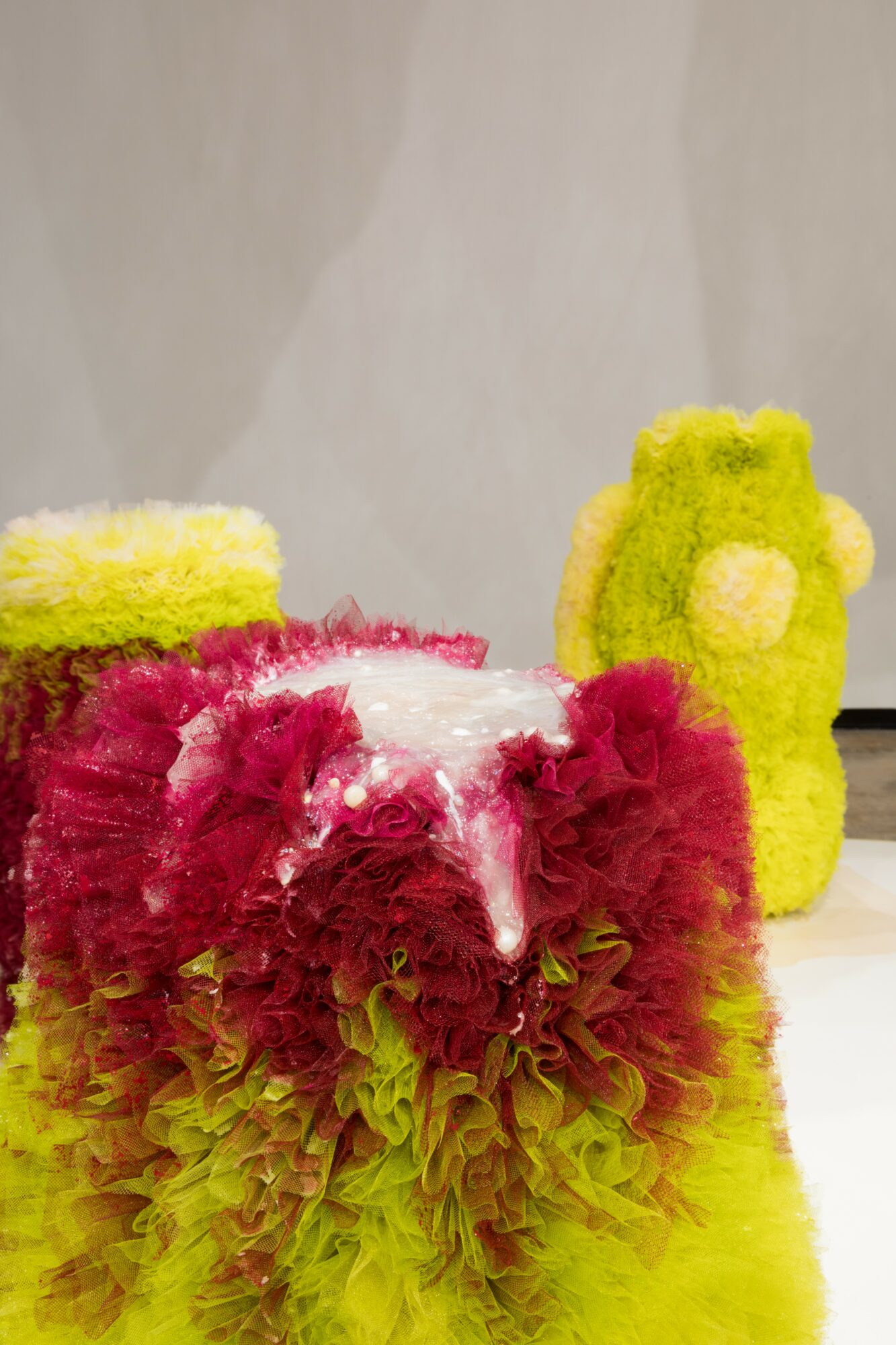 Colorful fuzzy toy with pink, yellow, and green sections, and a yellow plush figure in the background, on a white surface.
