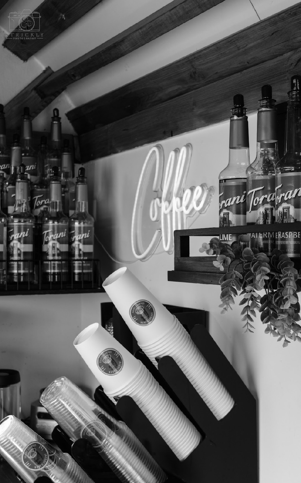 Coffee shop display with bottles, a neon sign, and cups on a counter, in black and white.