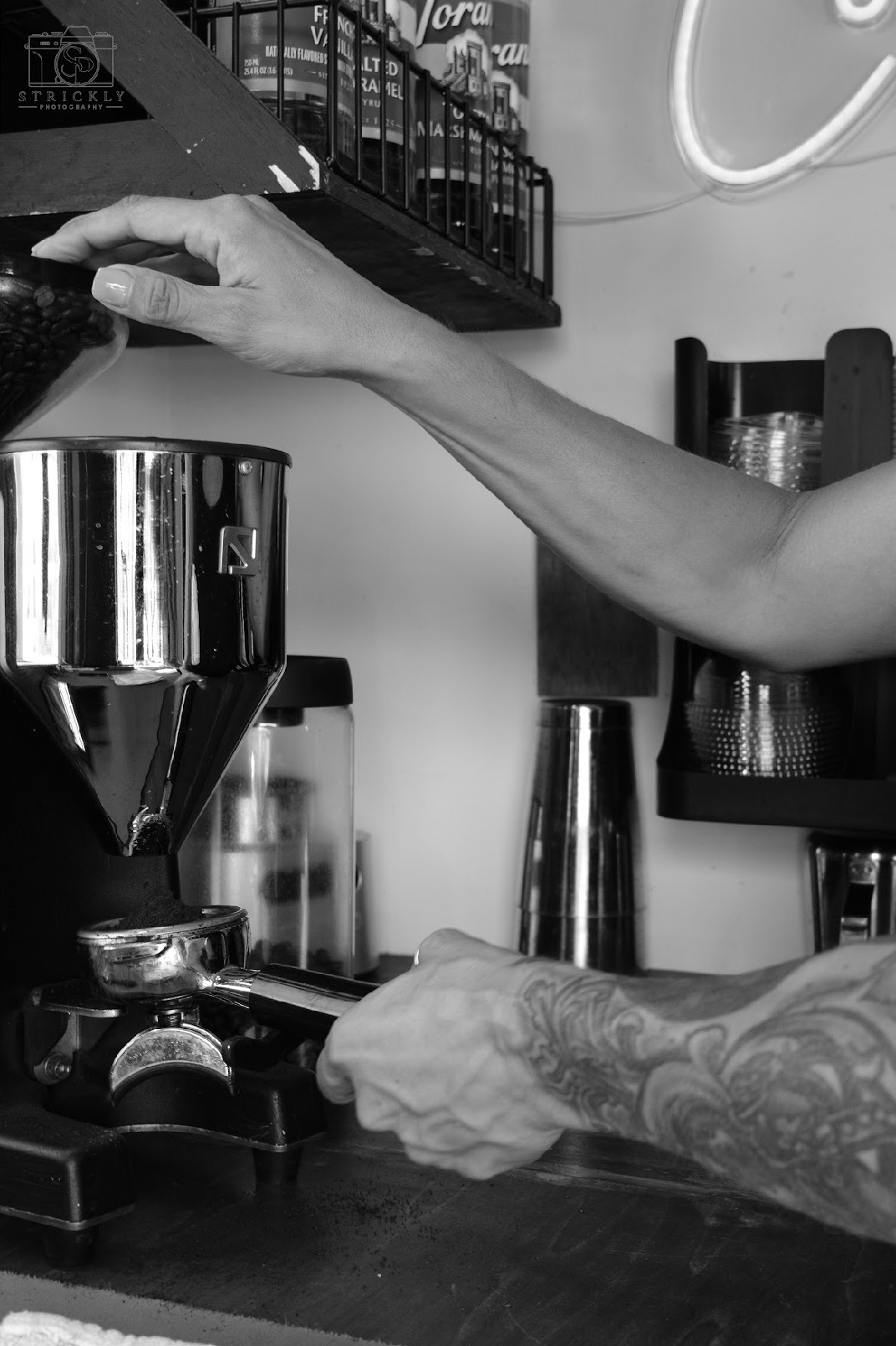 Person's arm and hand operating a coffee machine in a cafe, with various equipment and containers in the background.