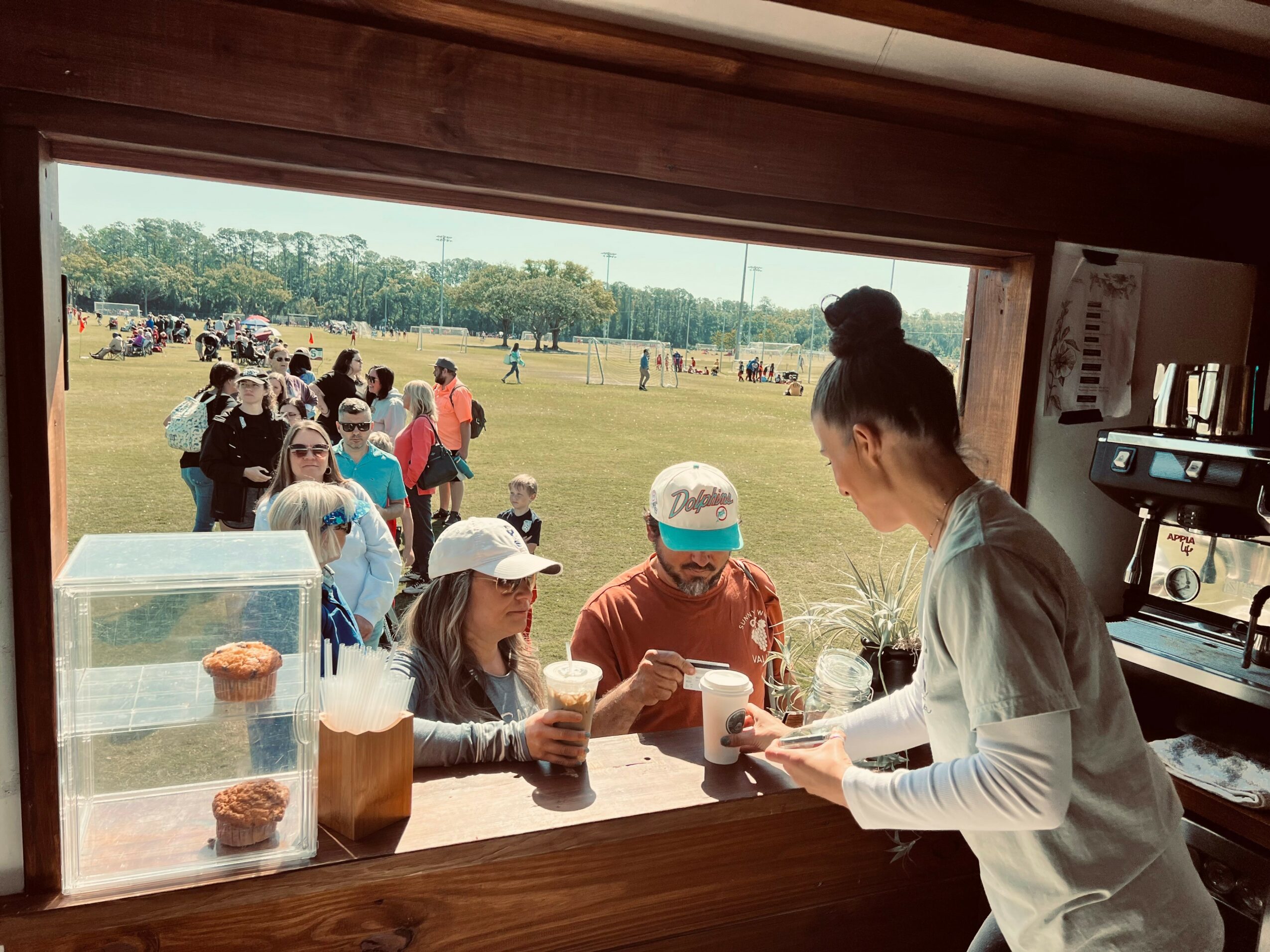 Person serving drinks to two children at a window, with a park and people outside, in a casual outdoor setting.