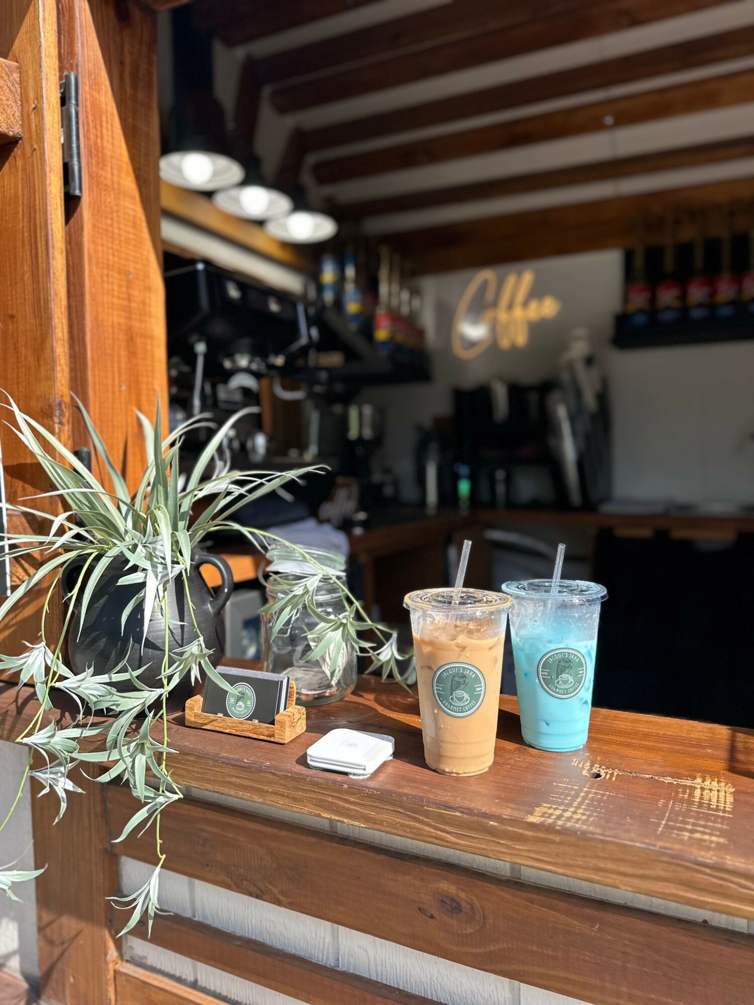 Two iced drinks on a wooden counter in a coffee shop, with a plant and a small chalkboard sign nearby.
