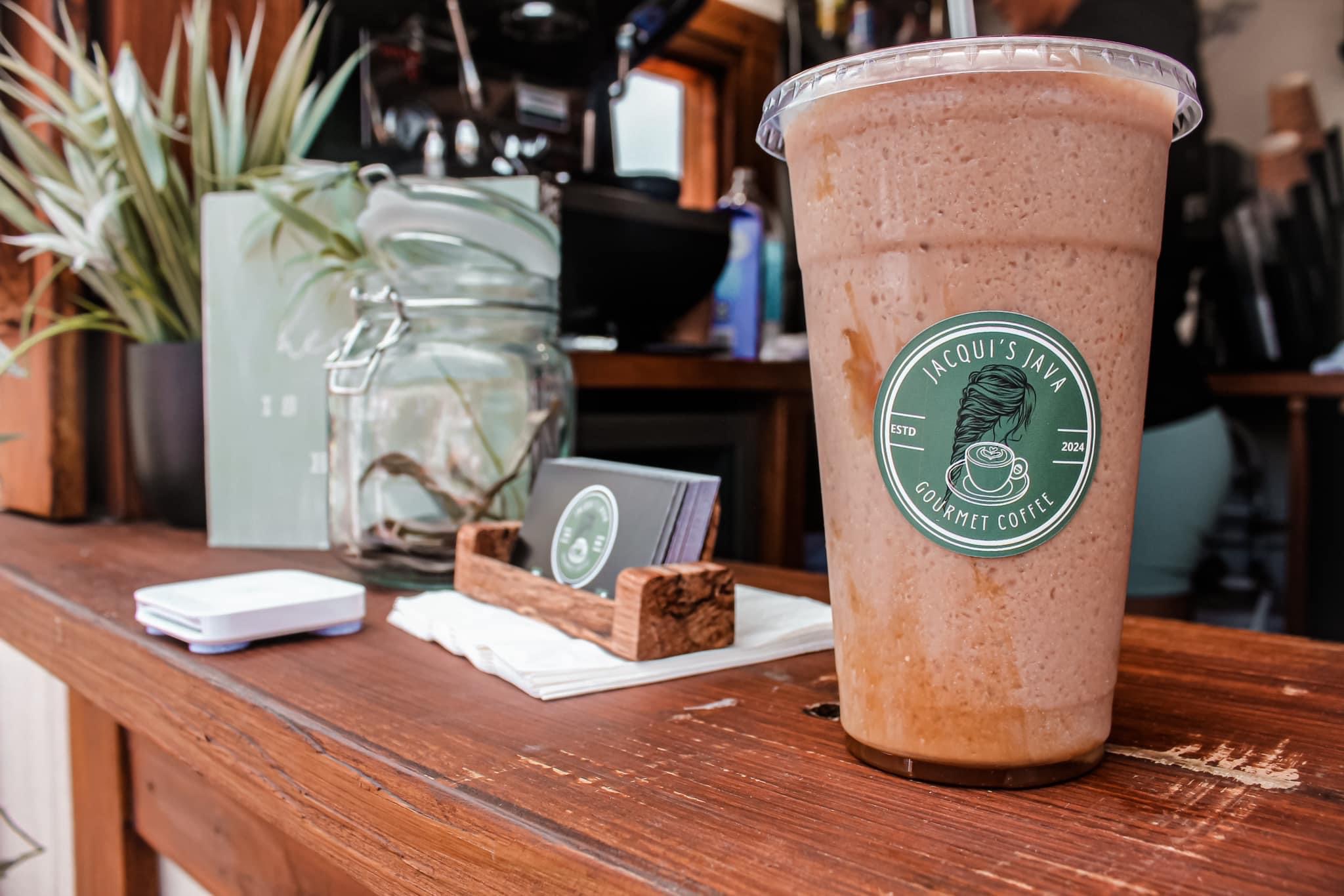 Large blended drink with a logo on a wooden table, with a plant and a glass jar in the background.