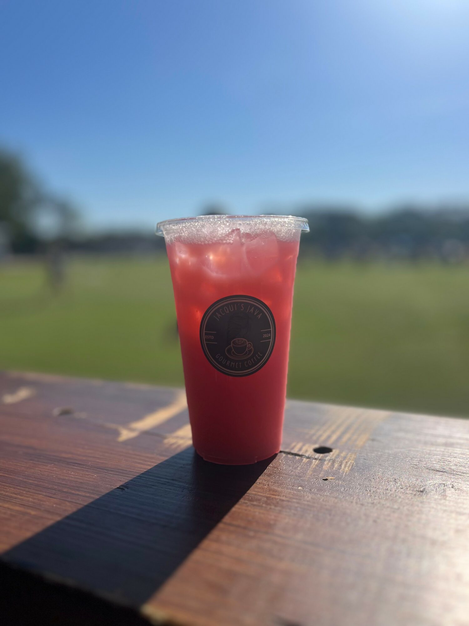 Red beverage in a clear cup with a black logo, outdoors on a sunny day, blurred green background, wooden surface