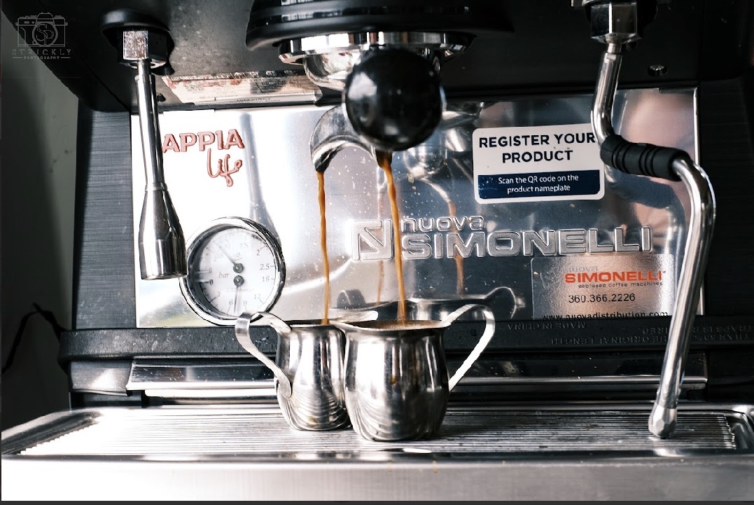 Close-up of a coffee machine pouring espresso into a small metal cup, with various labels and wires visible in the background.