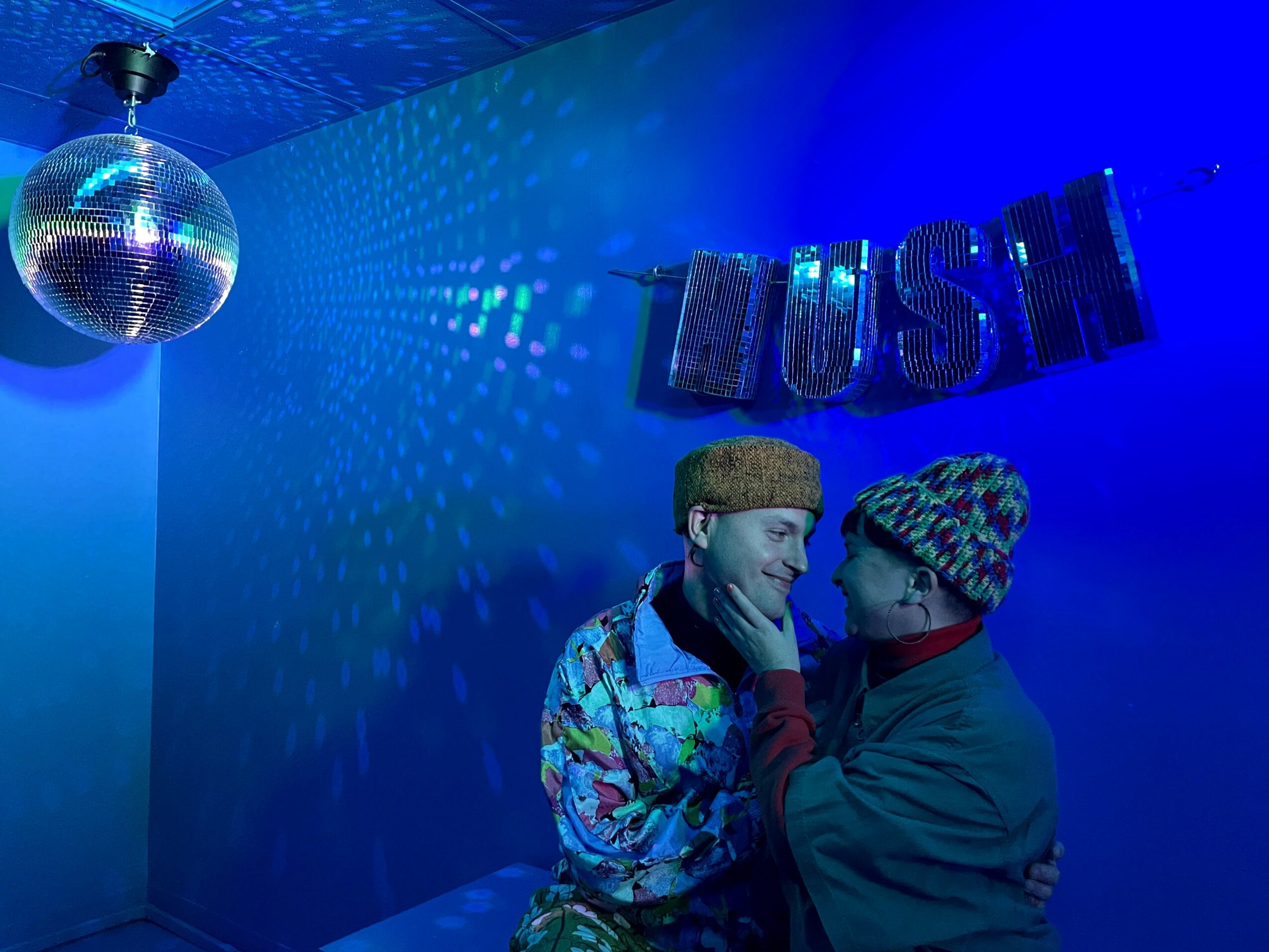 Two people in colorful clothing and hats close together in a blue-lit room with a disco ball and 'HUSTLE' sign.