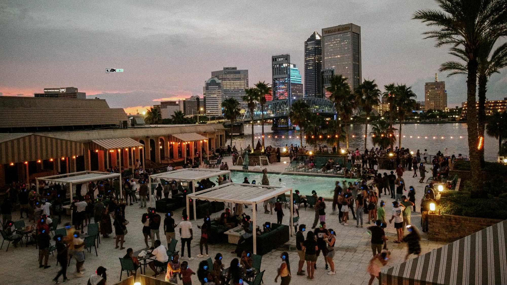 Crowd gathered at an outdoor event with city skyline and palm trees in the background during evening.