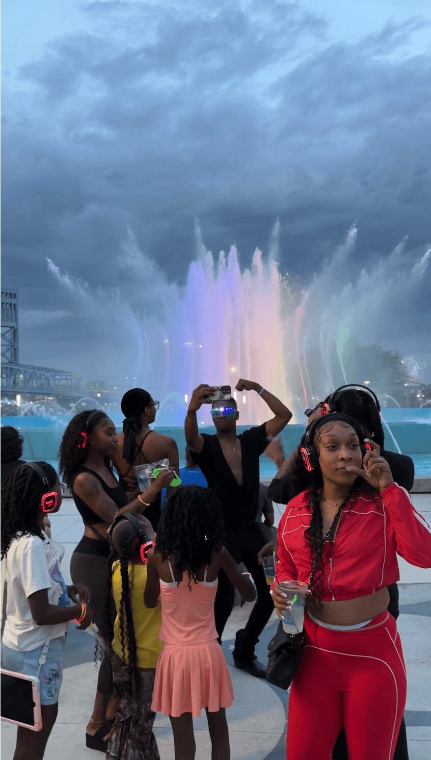 Group of children and adults watching a water fountain show outdoors under cloudy sky, some taking photos.