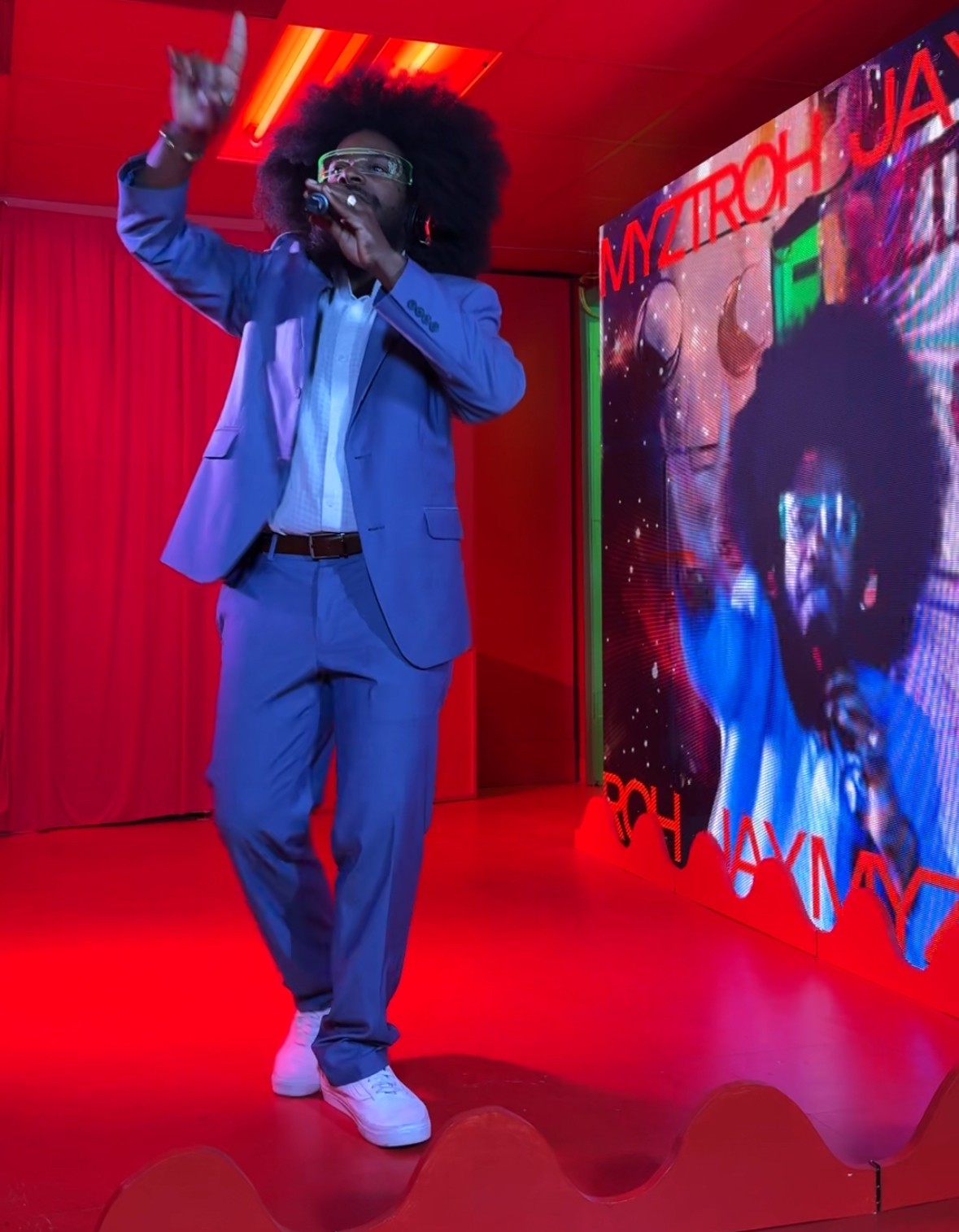 Person with curly hair in a blue suit singing or speaking on stage with a red curtain and a large screen behind.