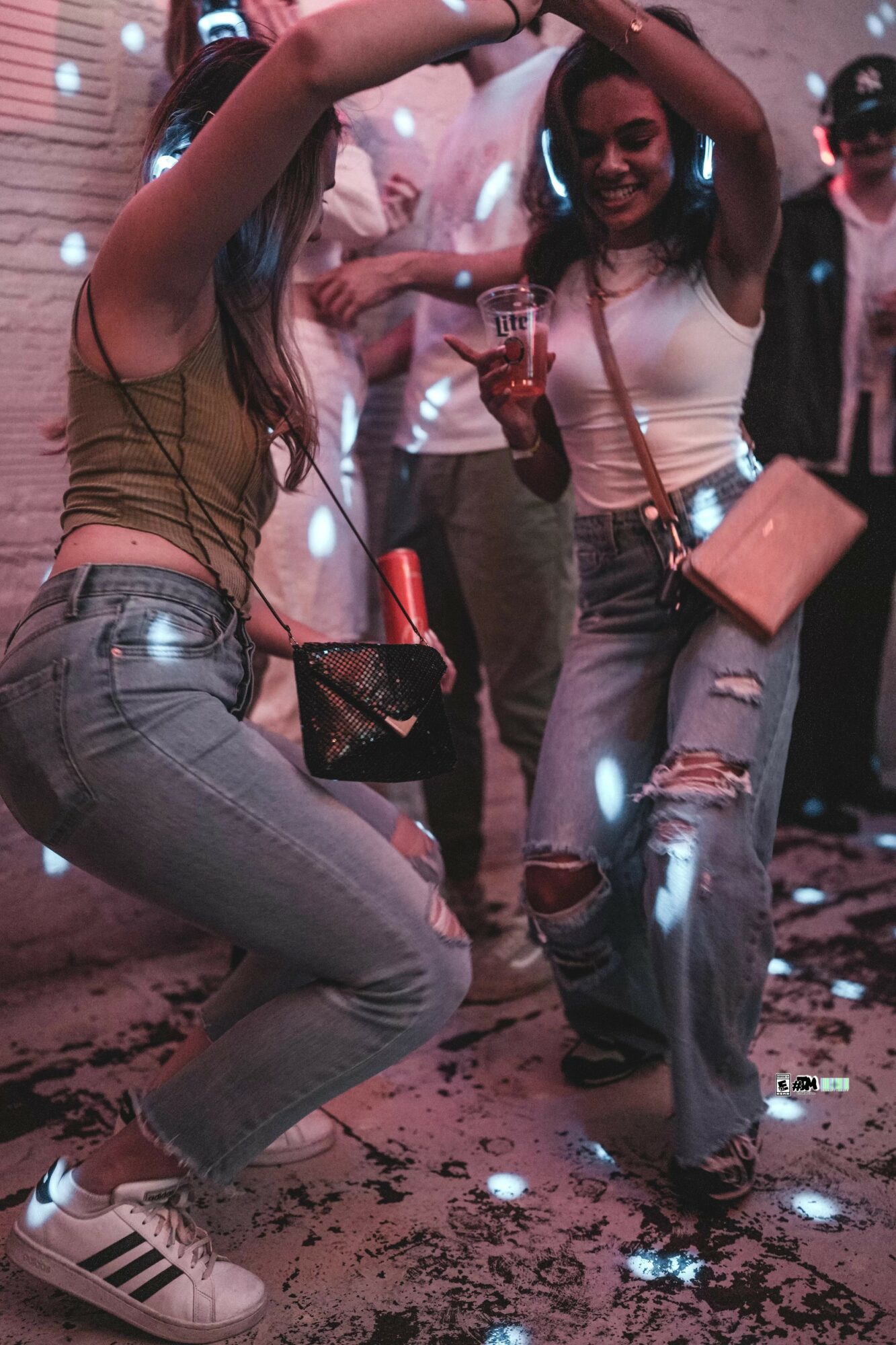 Two women dancing at a party, one holding a drink, with colorful lights and a crowd in the background.