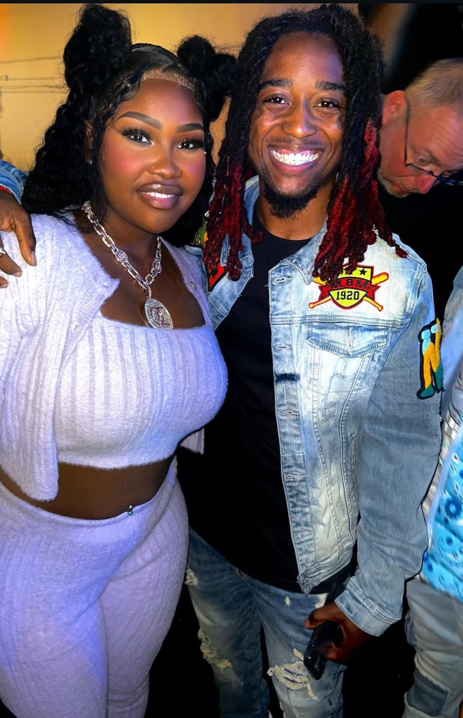 Two smiling people pose together, one wearing a white top and jewelry, the other in a denim jacket with patches.